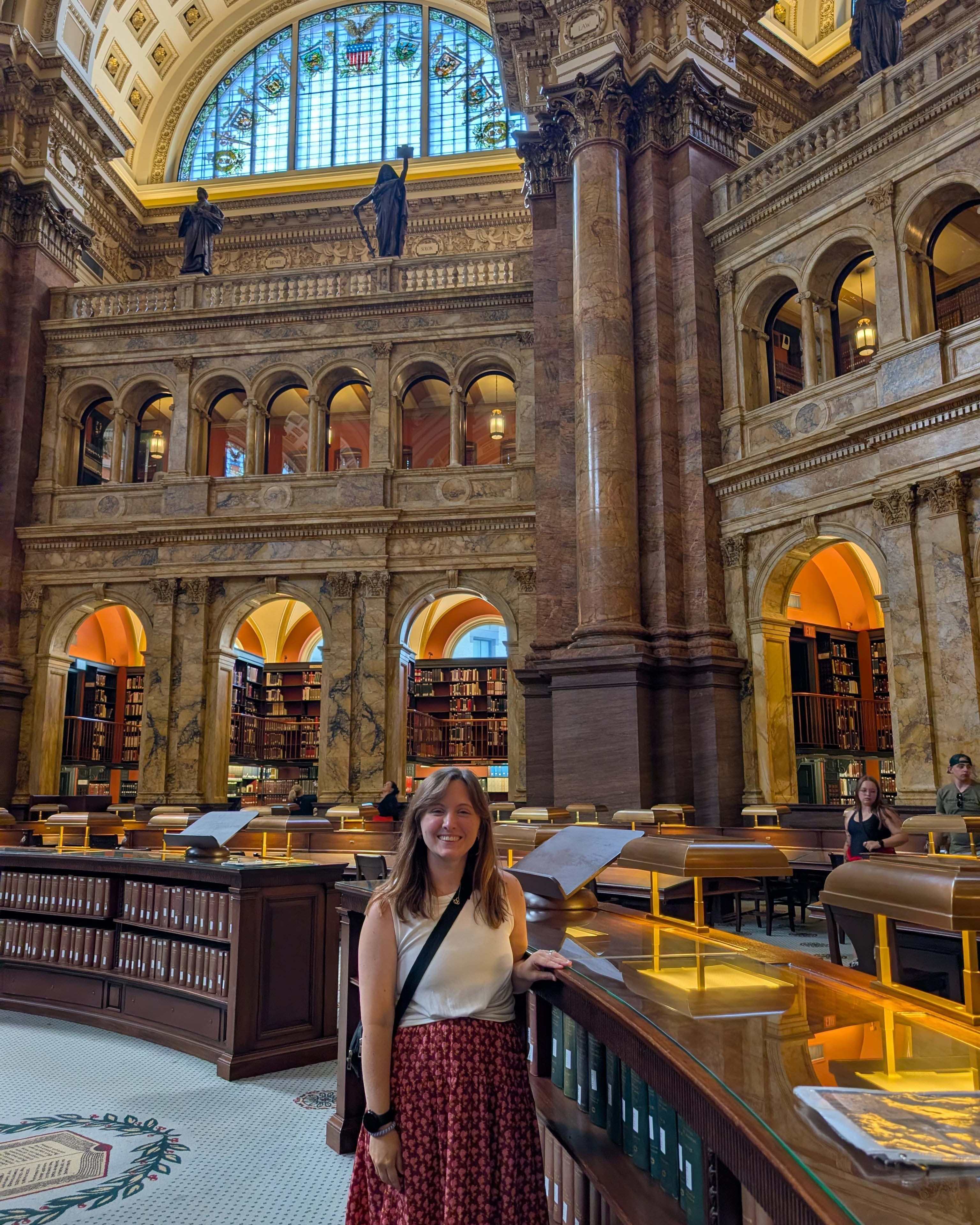 A woman stands in an ornate library with high ceilings, large columns, and arched windows. Bookshelves are on either side.