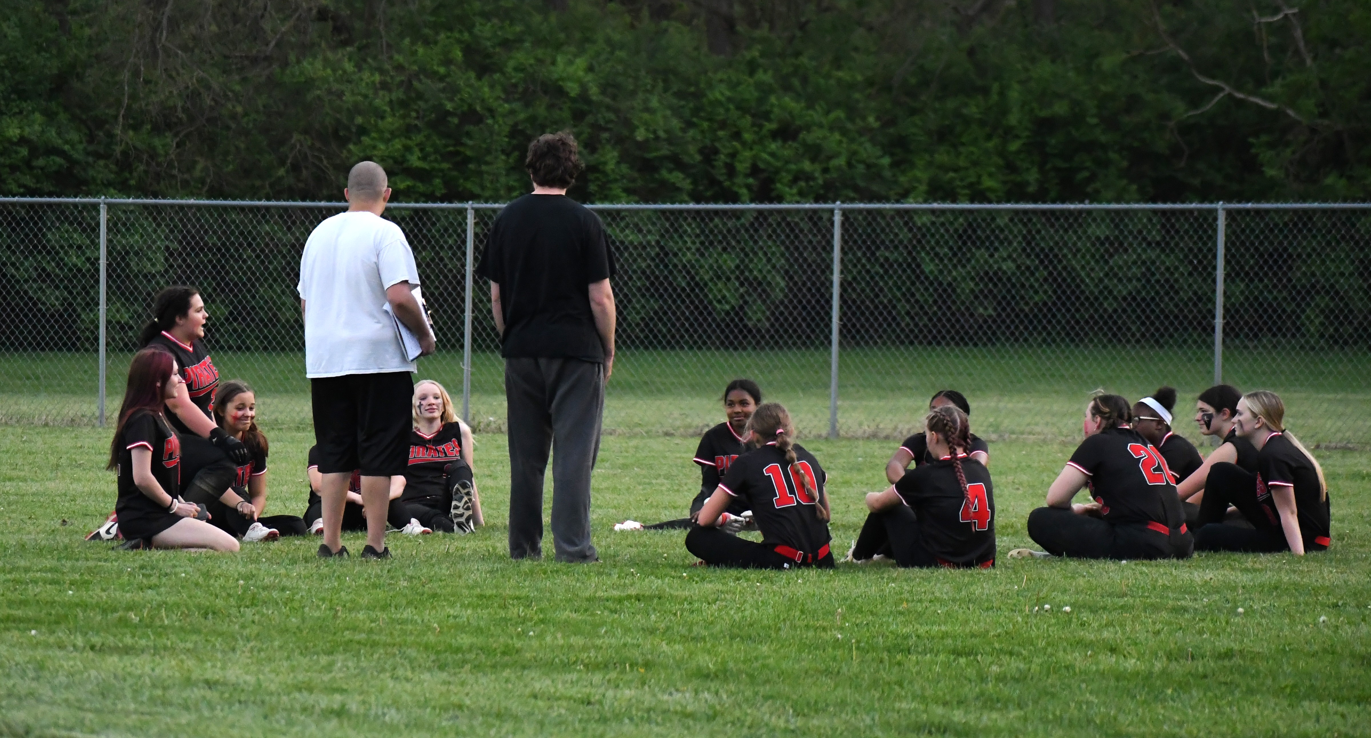 Softball players sitting in the grass with coaches speaking with them