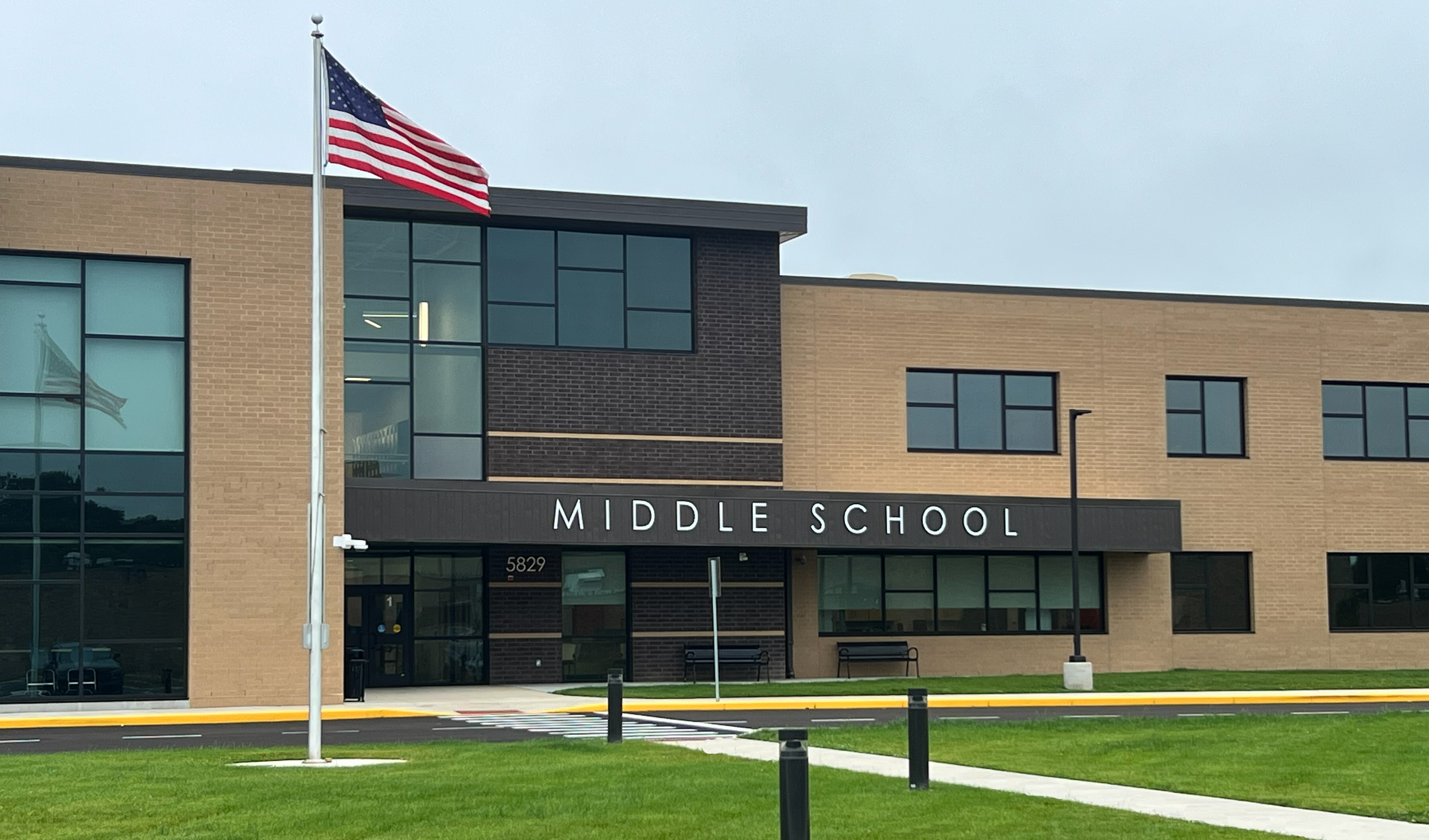 A middle school building with a flagpole displaying an American flag, large glass windows, and a grassy lawn.