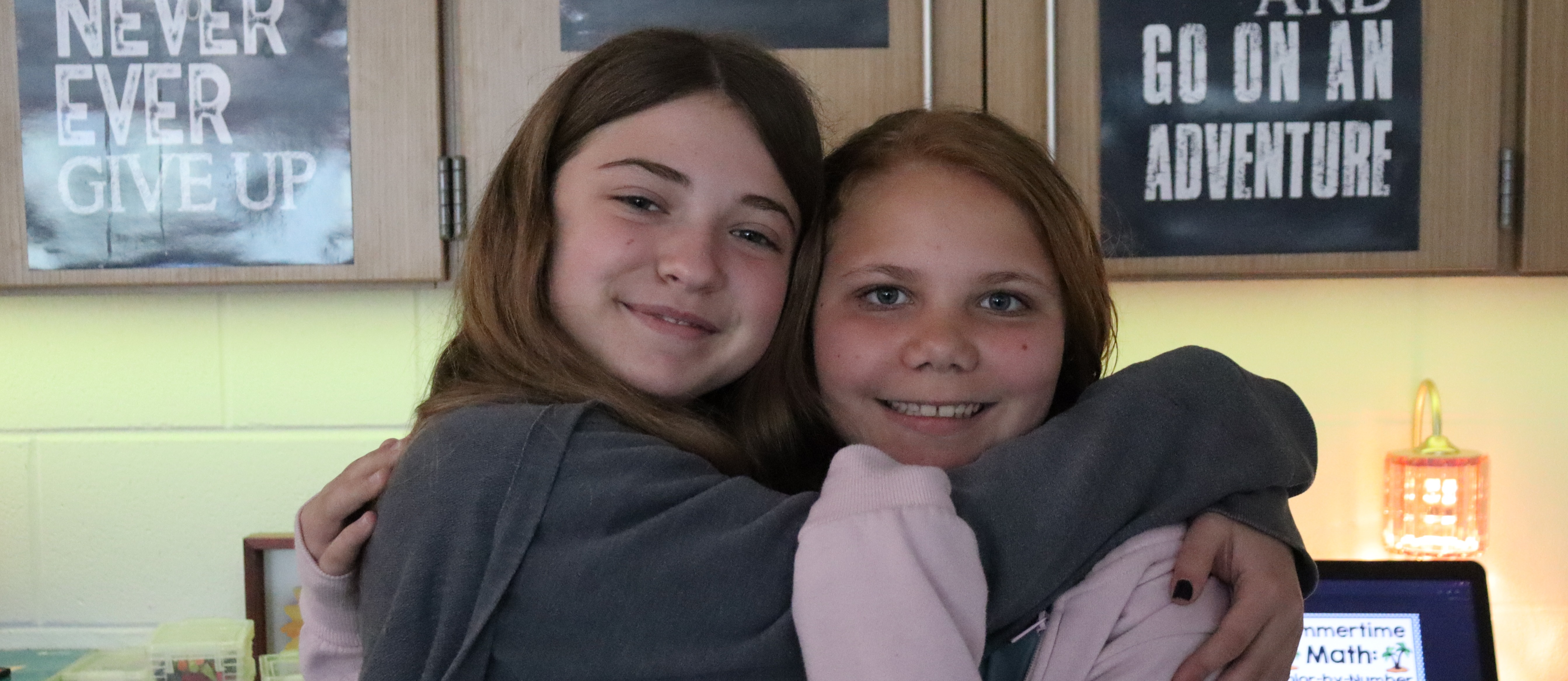 Two girls embracing, smiling, in a room with motivational posters. One wears a gray hoodie, the other a pink top.