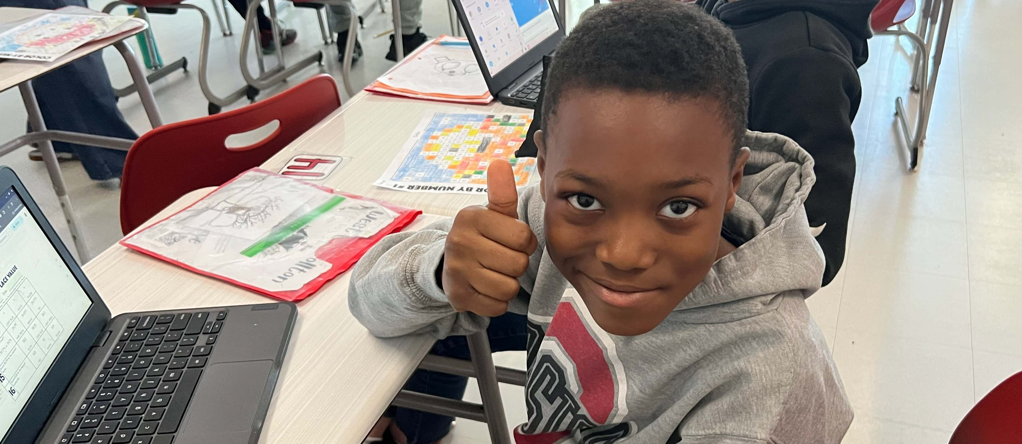 A young boy gives a thumbs-up, seated at a desk with a laptop in a classroom.