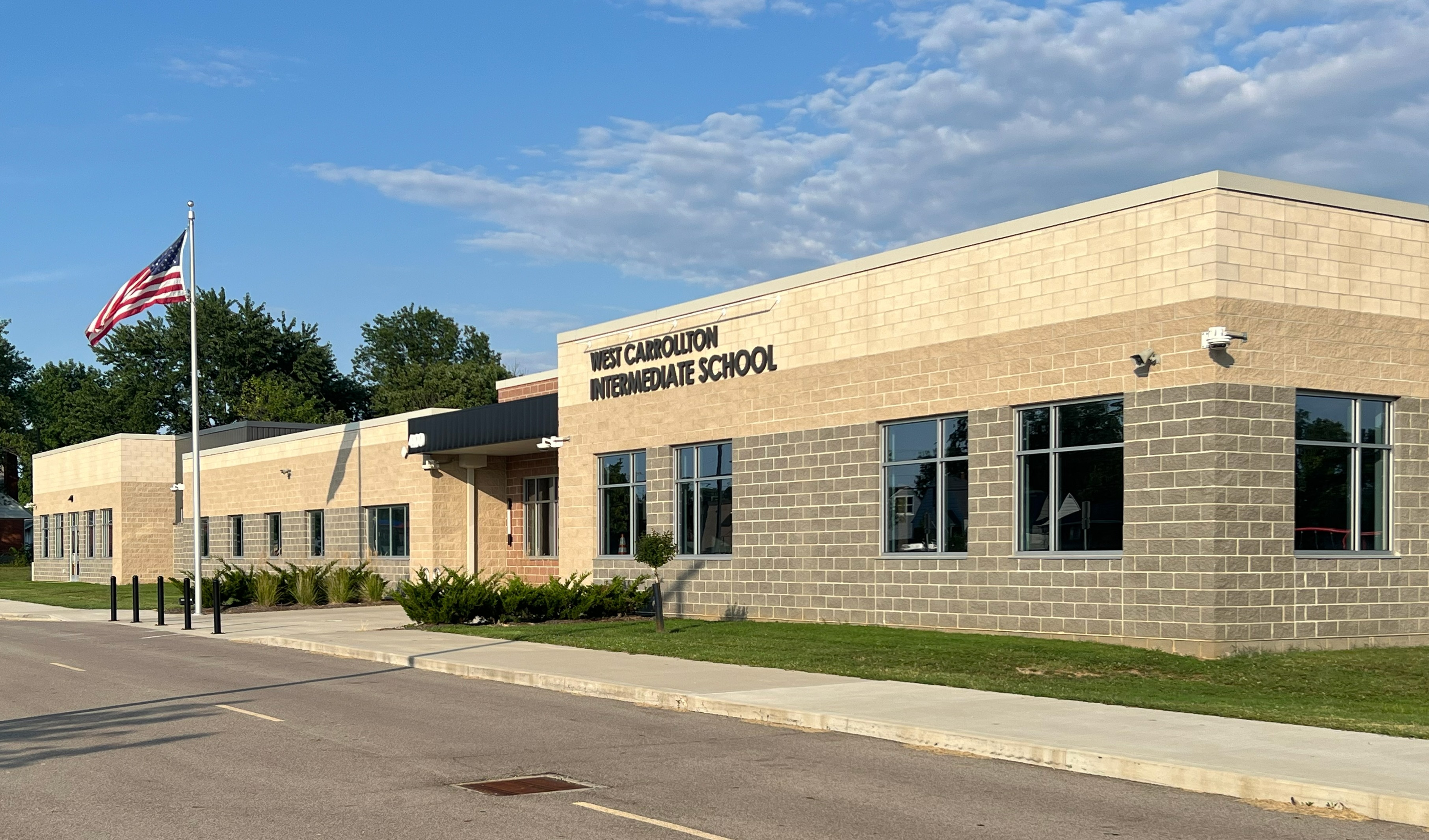 A modern school building with brick walls and glass windows. An American flag flies from a flagpole. The sidewalk and road are empty.