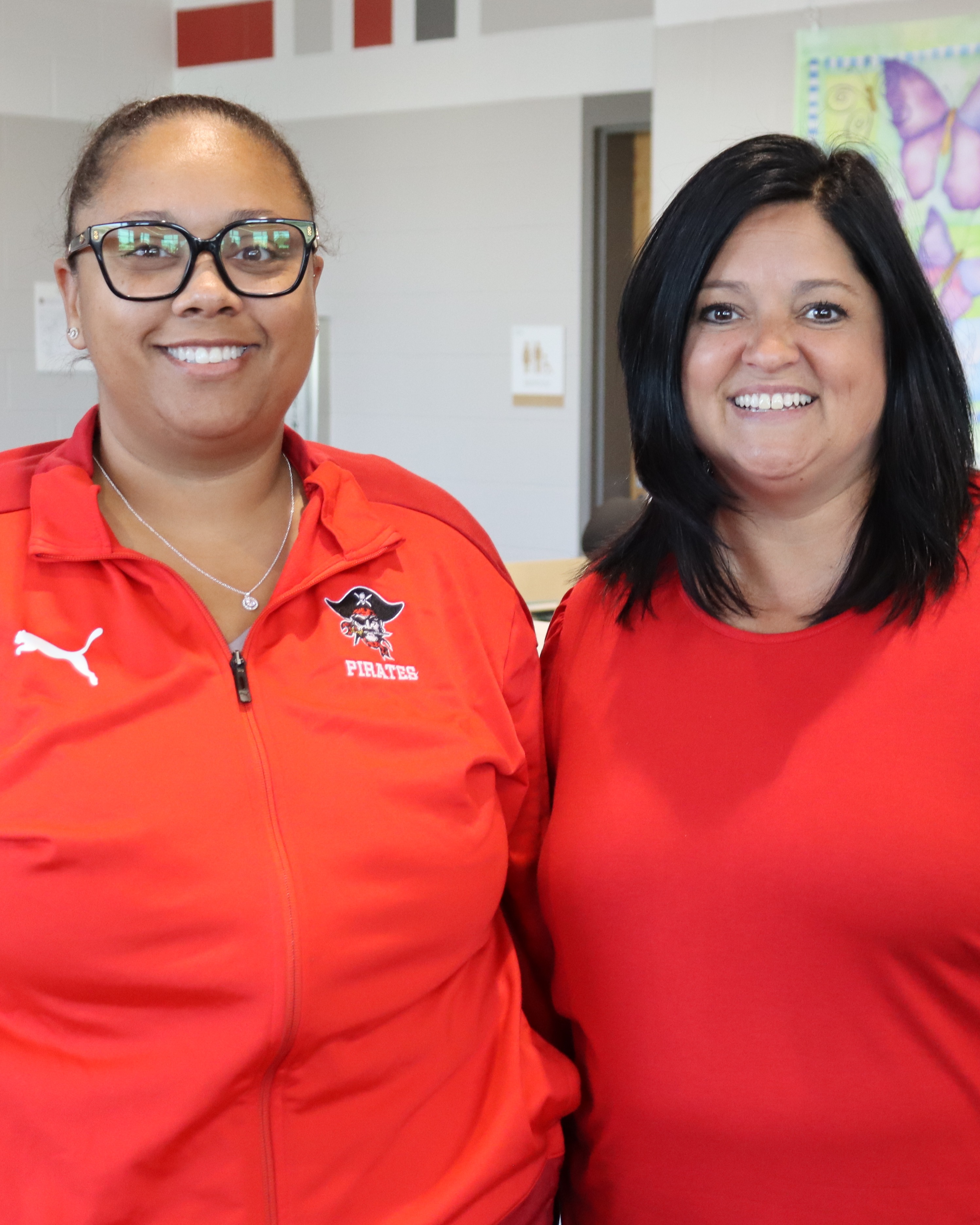 Two women in red shirts, one with glasses, standing next to each other in an indoor office.