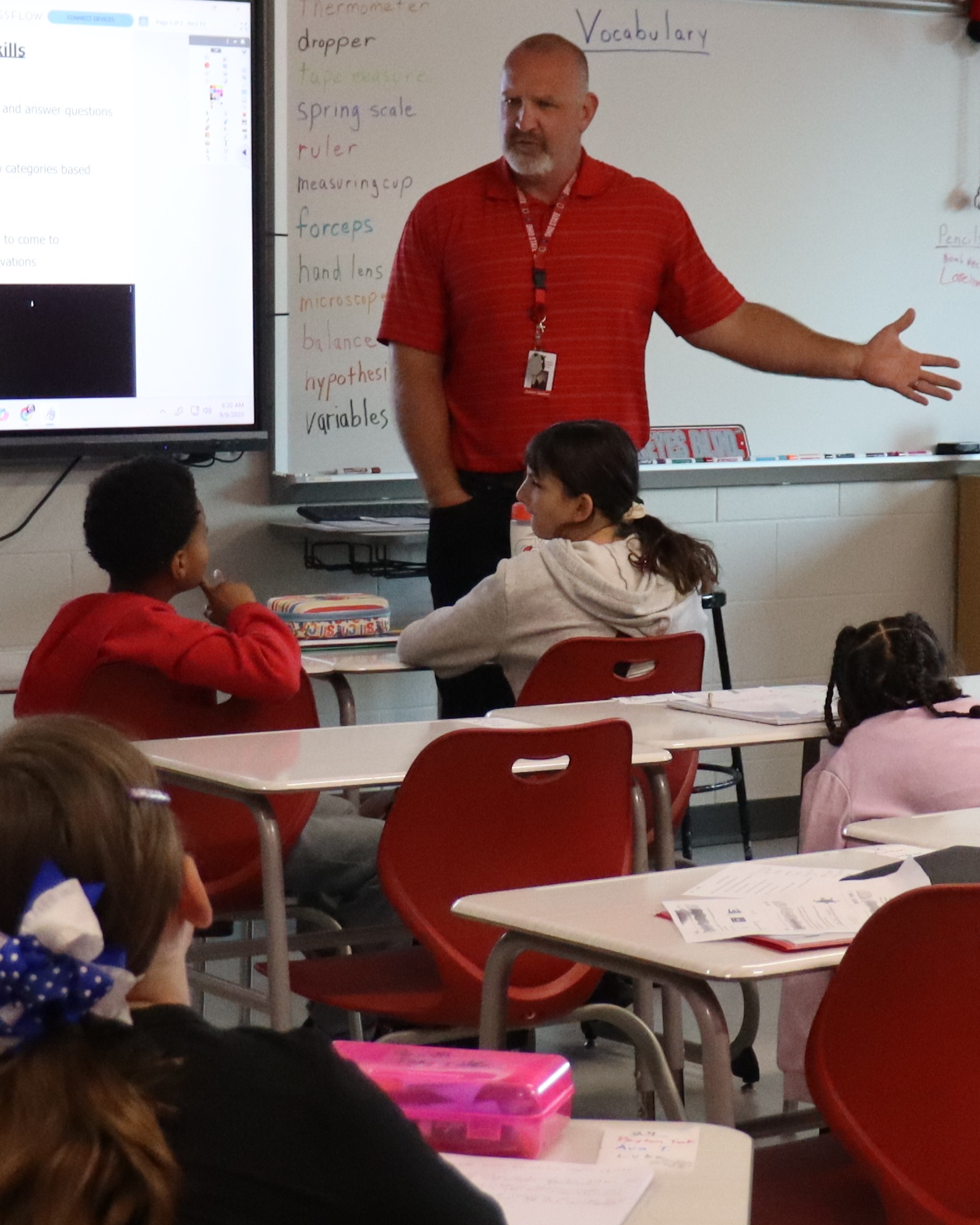 A classroom with students seated at desks. A teacher stands before a whiteboard and screen, presenting to the class.