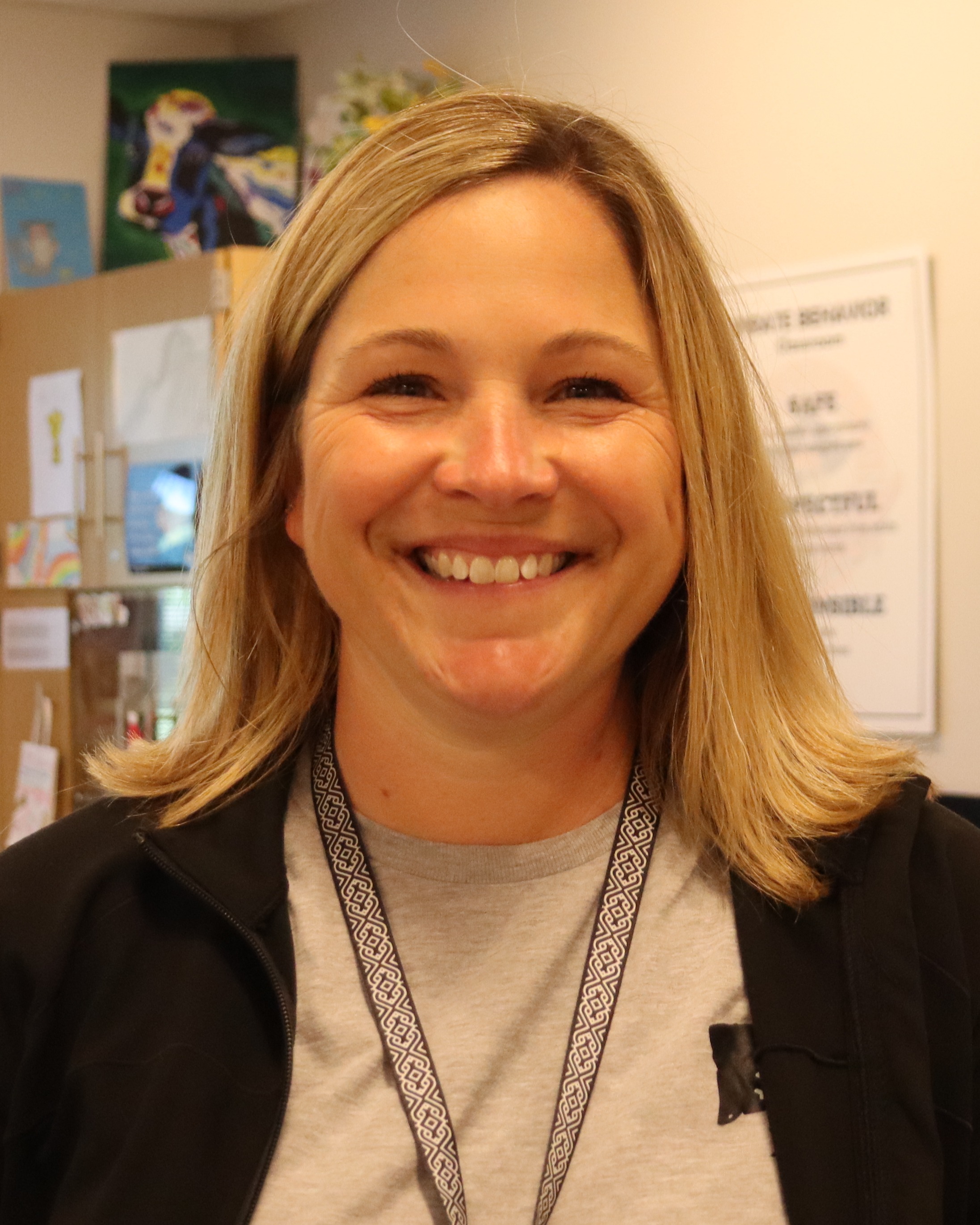 A smiling woman with blonde hair stands in an office. She wears a gray shirt and a lanyard. Behind her, there's a whiteboard, plants, and a window with curtains.