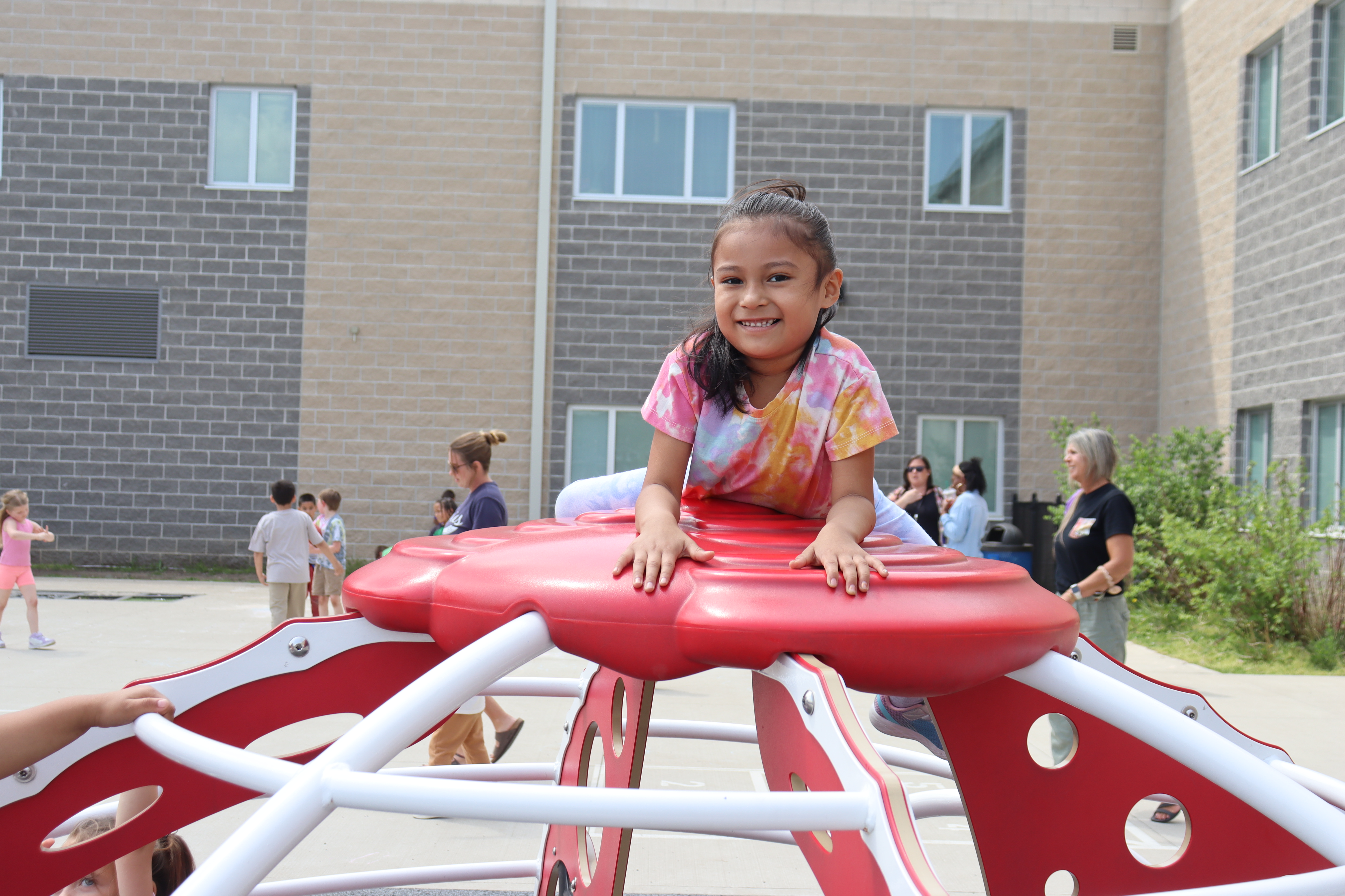 Girl in tie-dye shirt sitting on a red climbing structure with a building in the background.