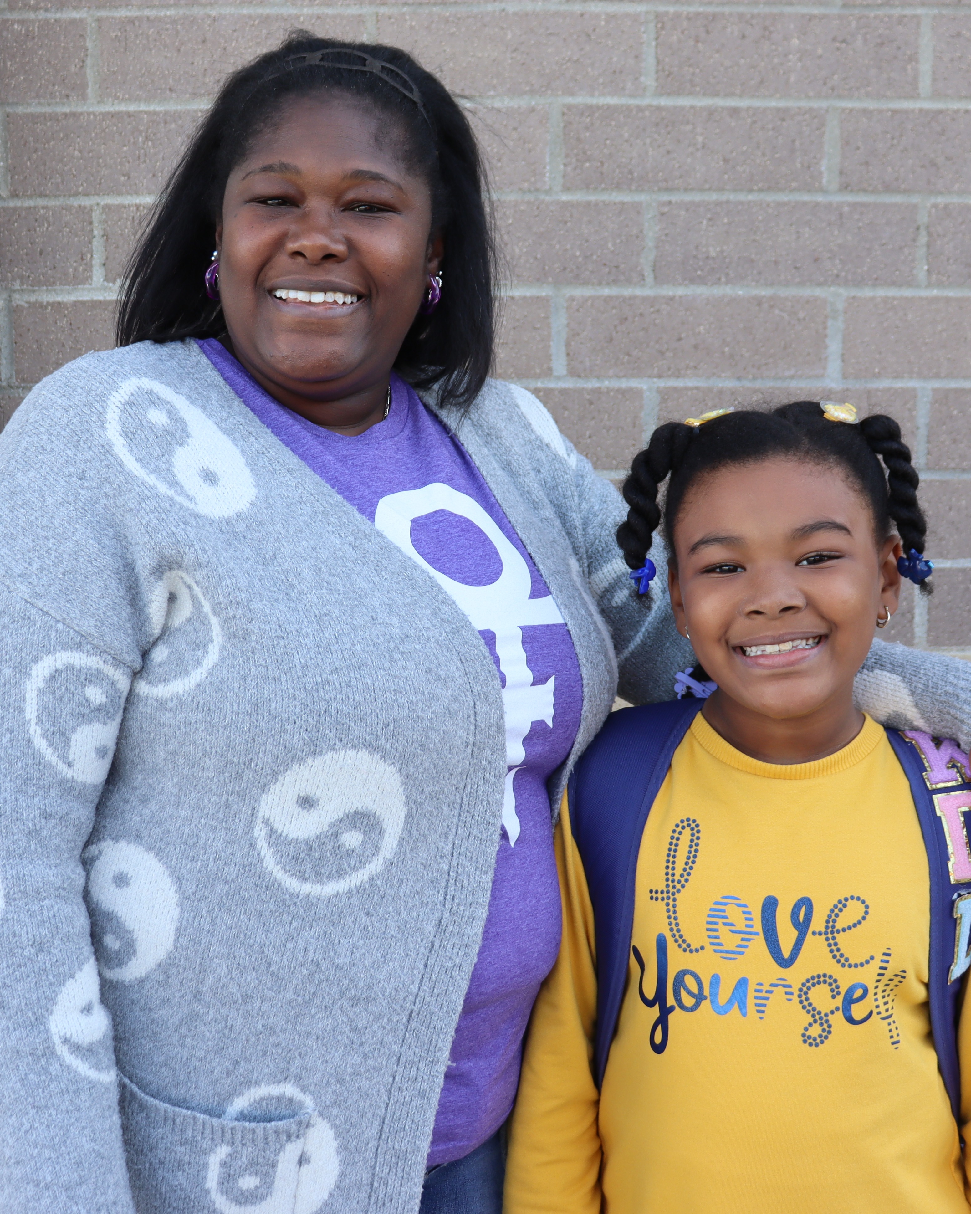 A smiling adult stands with a young girl, both wearing backpacks, in front of a brick wall.