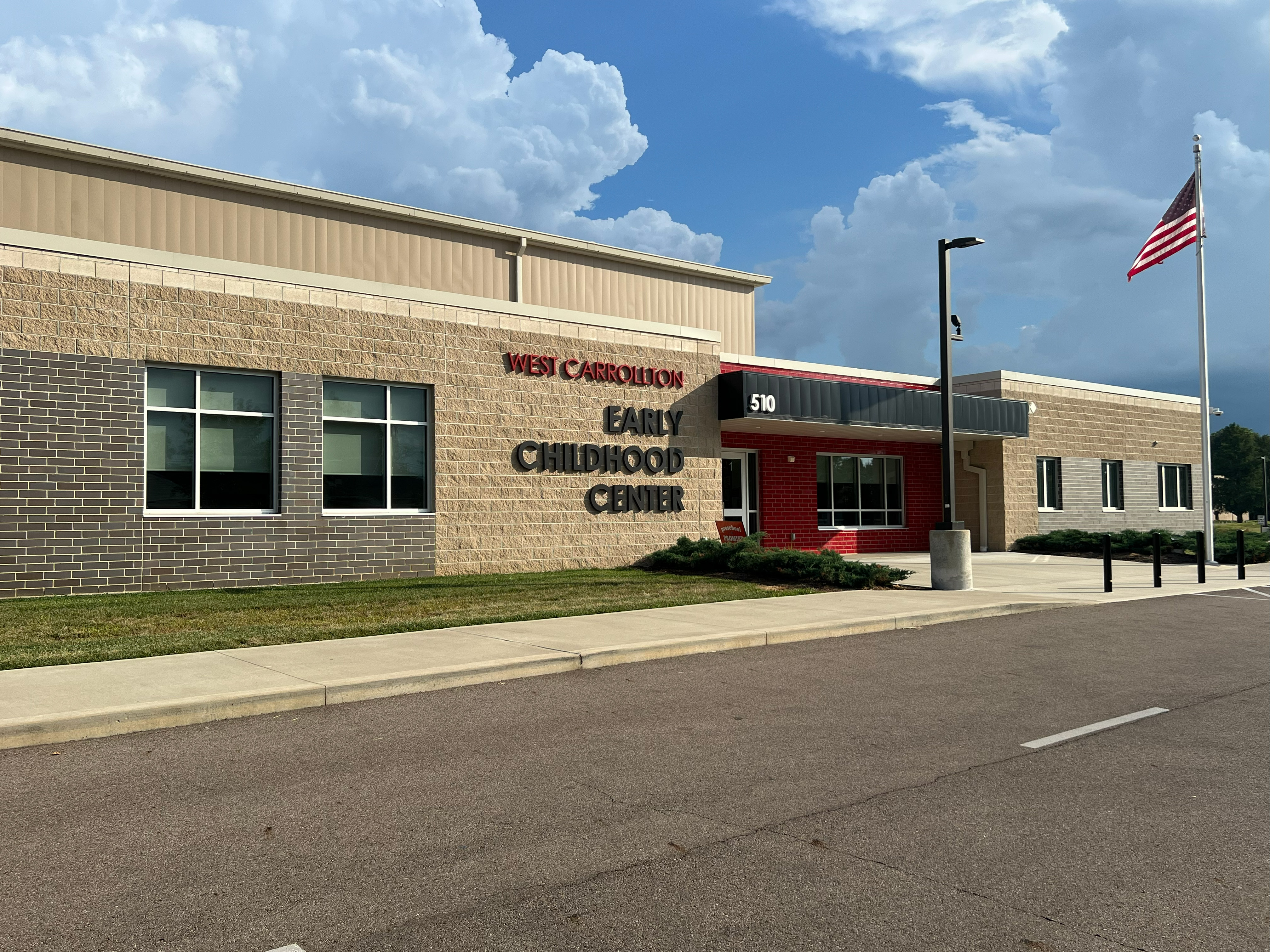 School building with light brown brick and a red entry way. A driveway is in the front of the image. In front of the building is a grassy area, sidewalk, and flag pole with an American flag. The sign on the building says West Carrollton Early Childhood Center