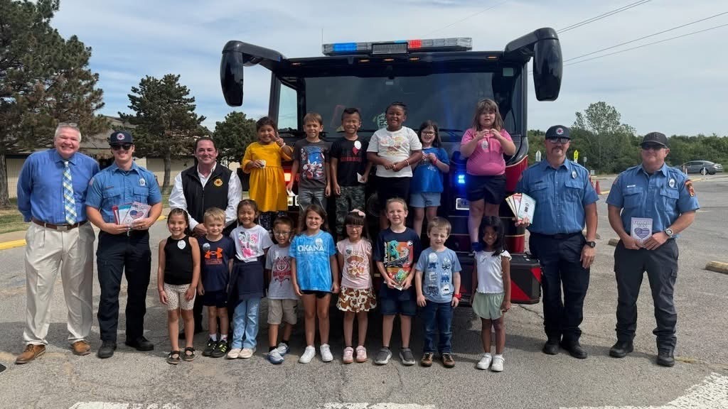Group of children and adults pose in front of a fire truck, smiling, on a sunny day.