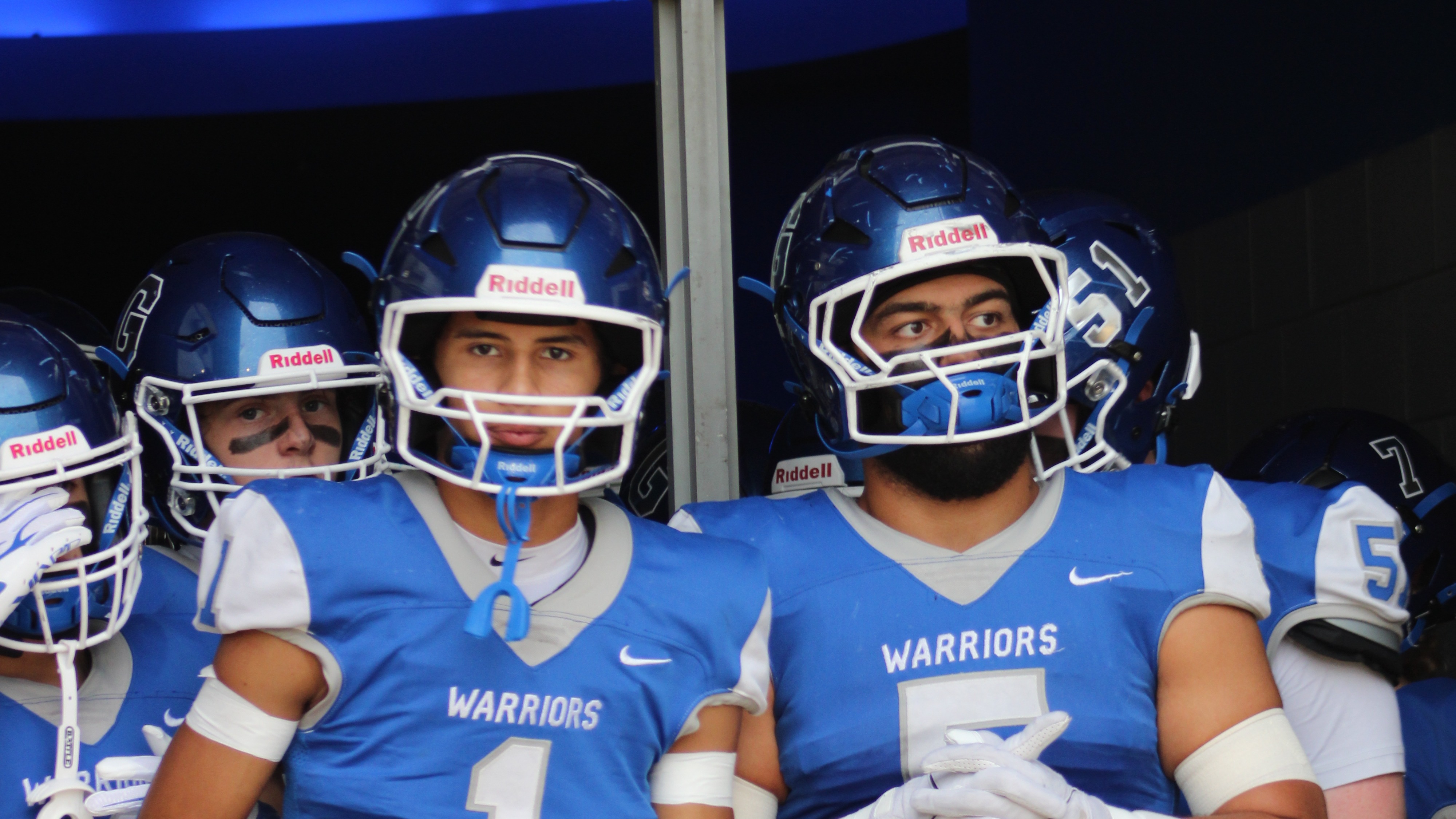 Athletes in blue and gray uniforms with numbers 1 and 5 stand in a locker room. They wear helmets and gloves.