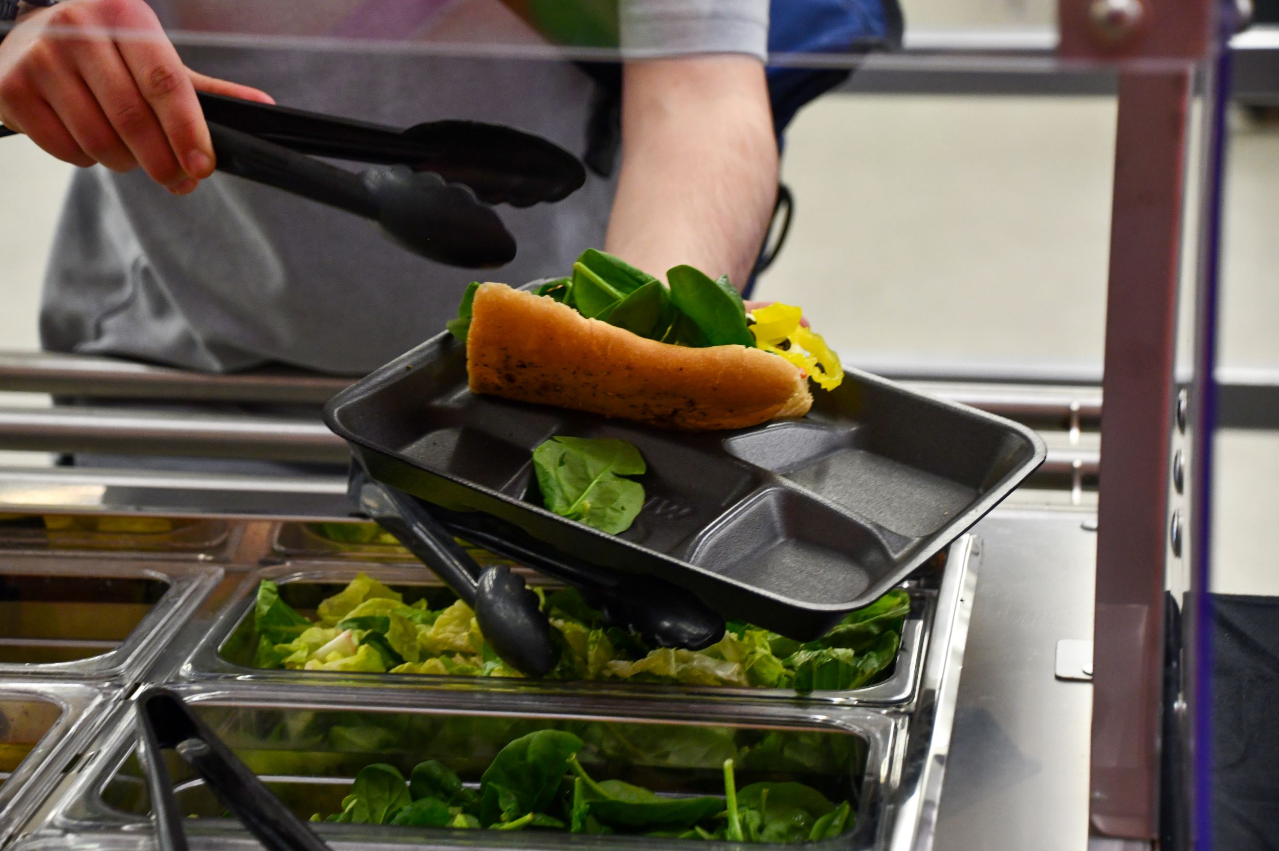 hand holding tongs and putting spinach on a sub sandwich