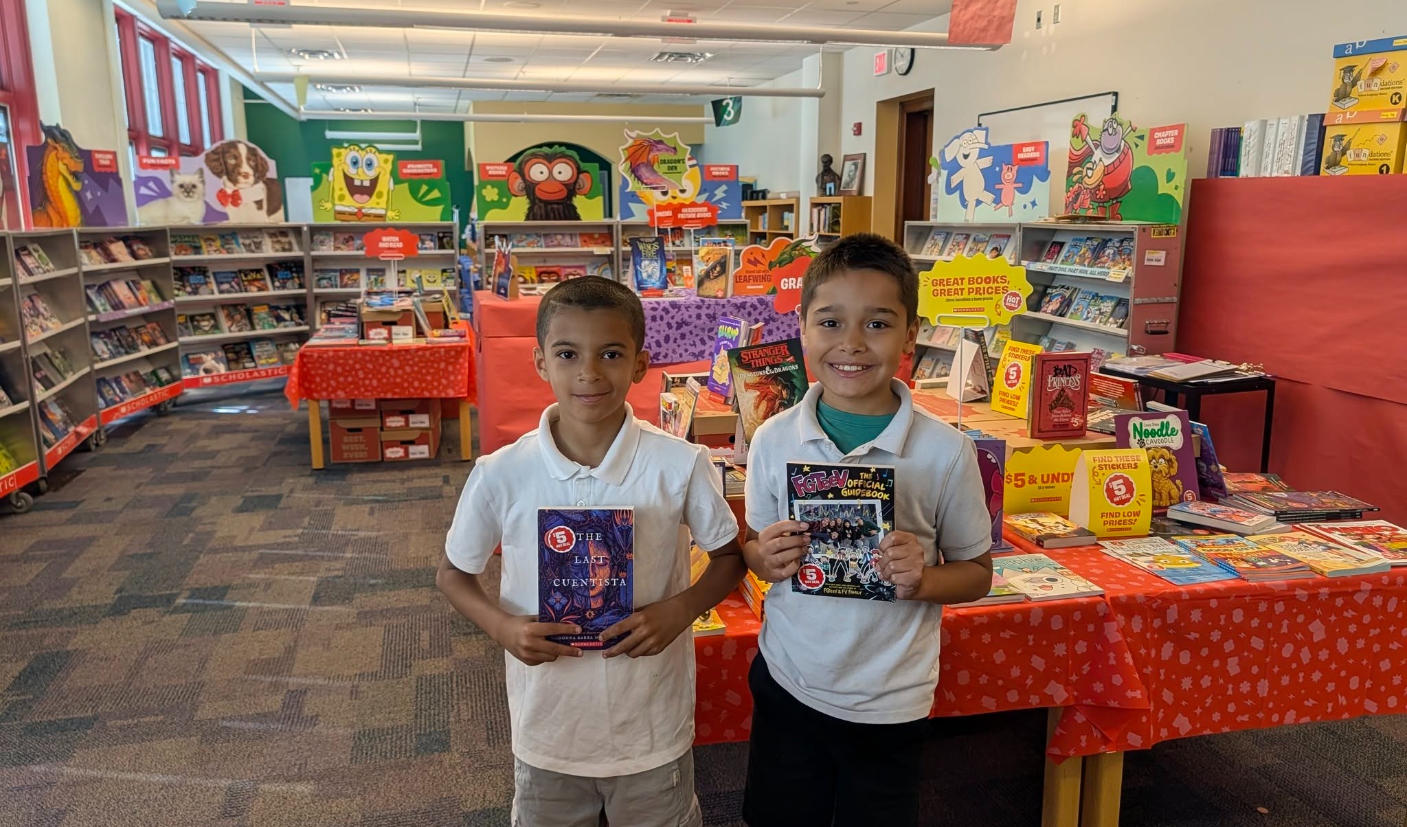 Two students are holding books at a colorful school book fair surrounded by displays and shelves of books.