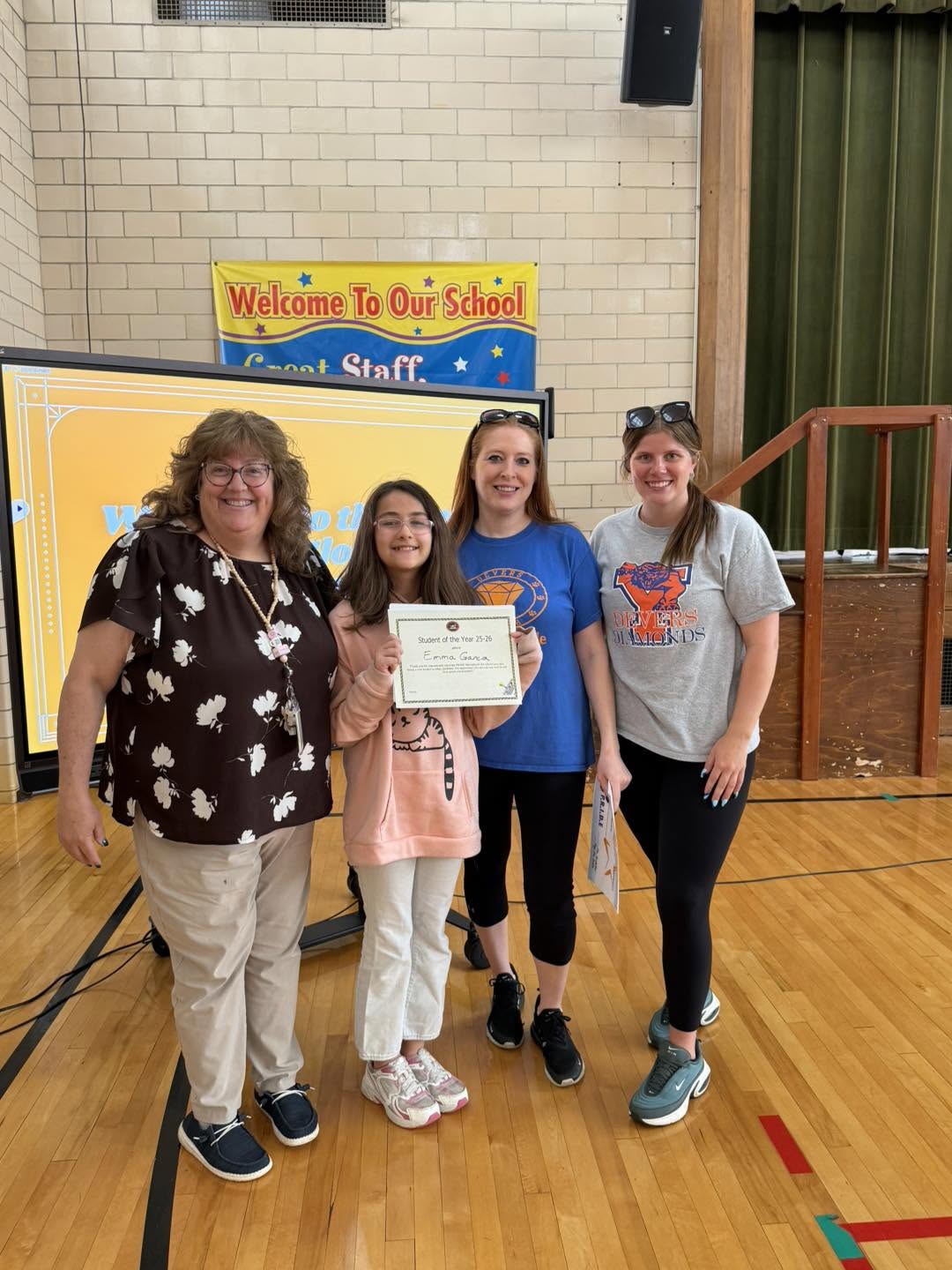 A student is holding a certificate and posing with three staff members in a school gym during an awards ceremony.