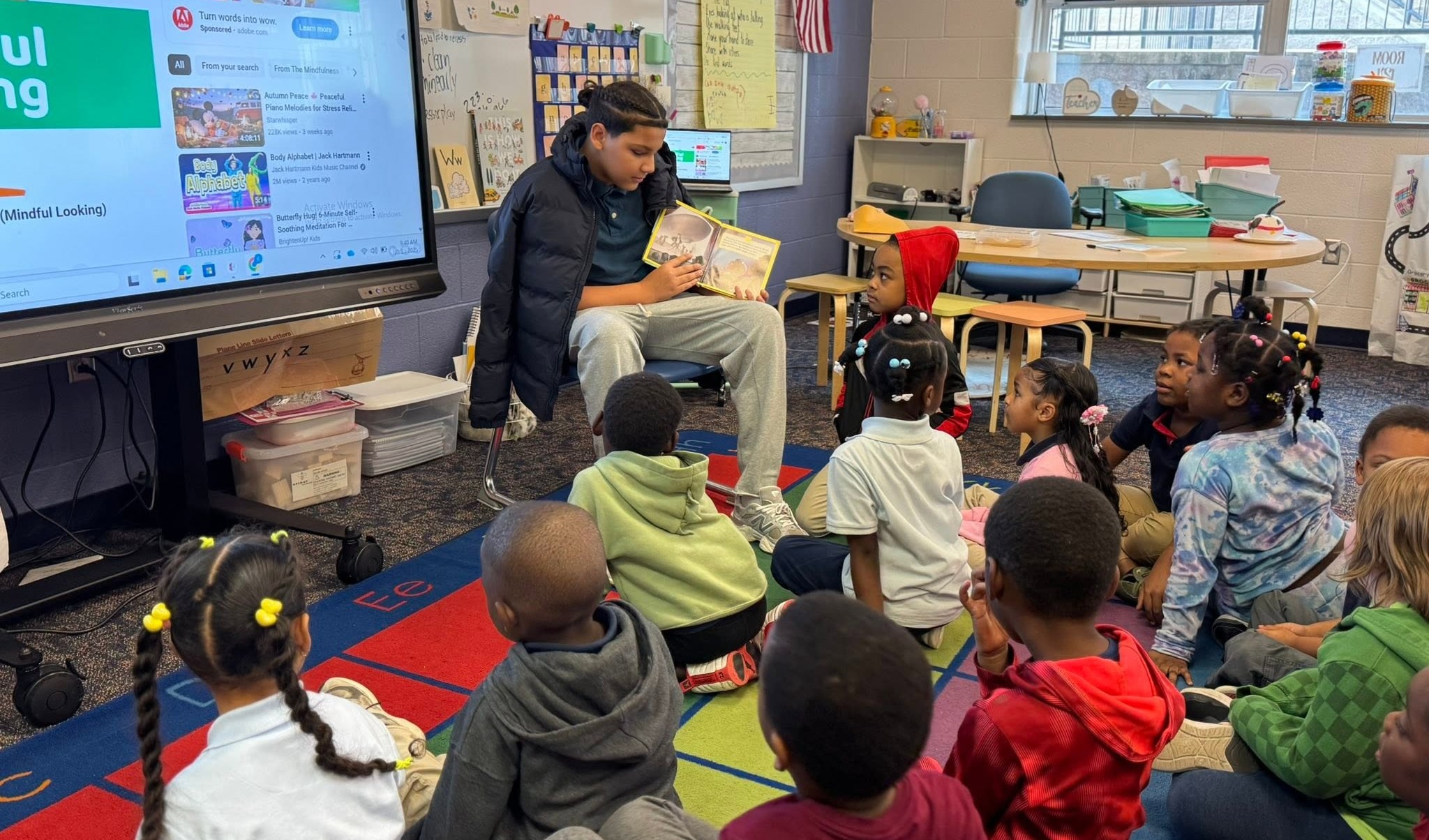 An older student is reading a book aloud to a group of younger students who are seated on a school classroom rug.
