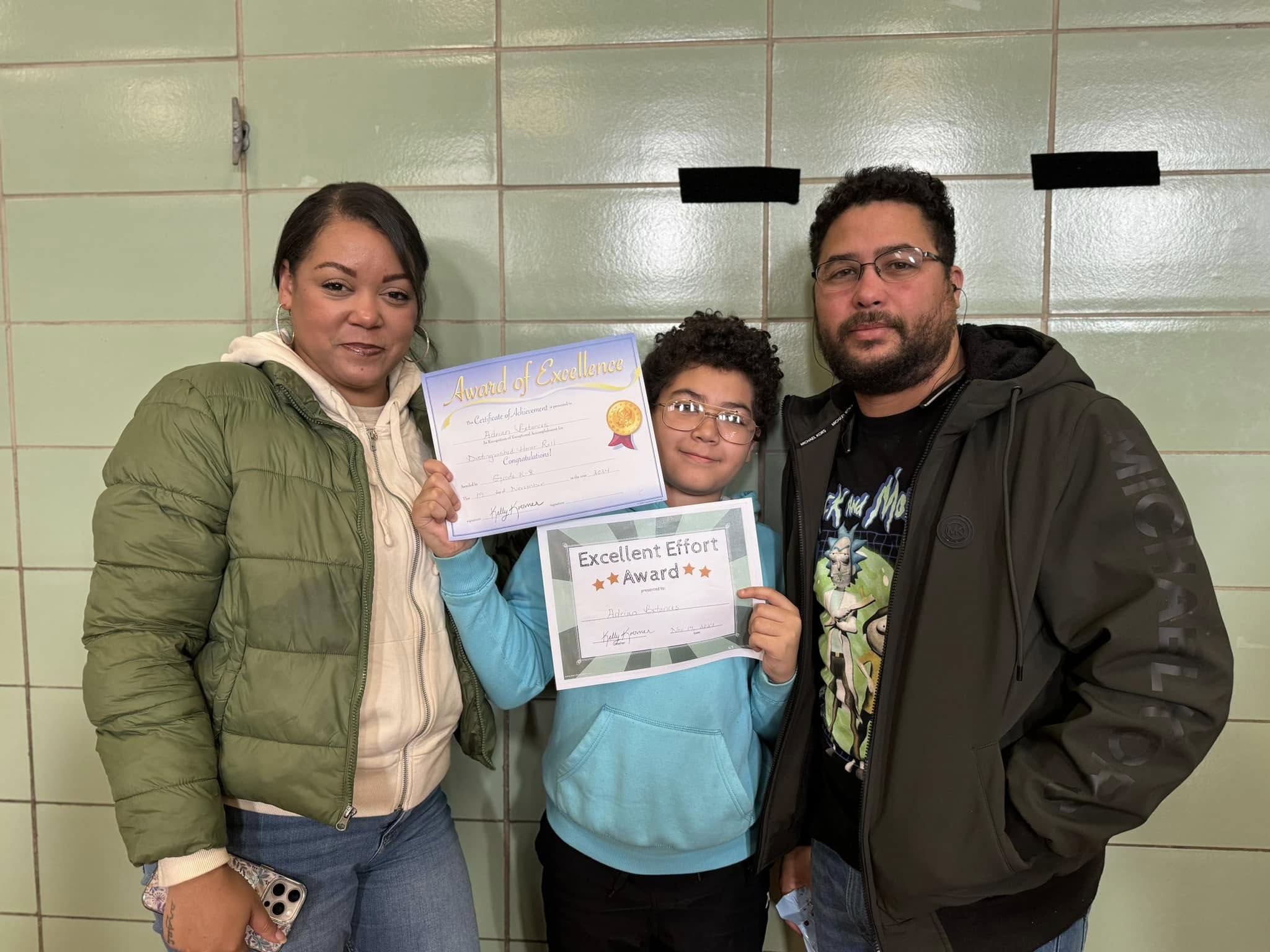 A child stands between two adults, holding two certificates: one for “Award of Excellence” and another for “Excellent Effort Award.” All three are smiling, and they are standing against a wall with green tiles and black tape behind them.