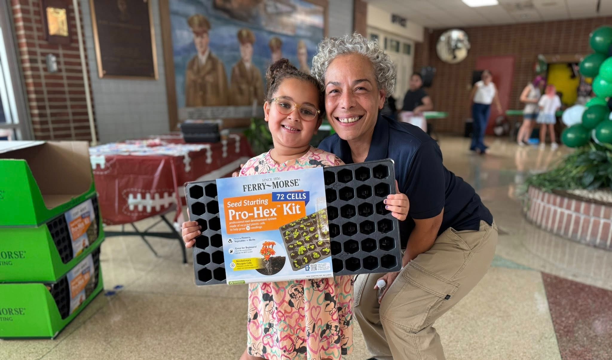 A district staff member bending down next to a student holding up a book titled "Seed Starting Pro-Hex Kit."