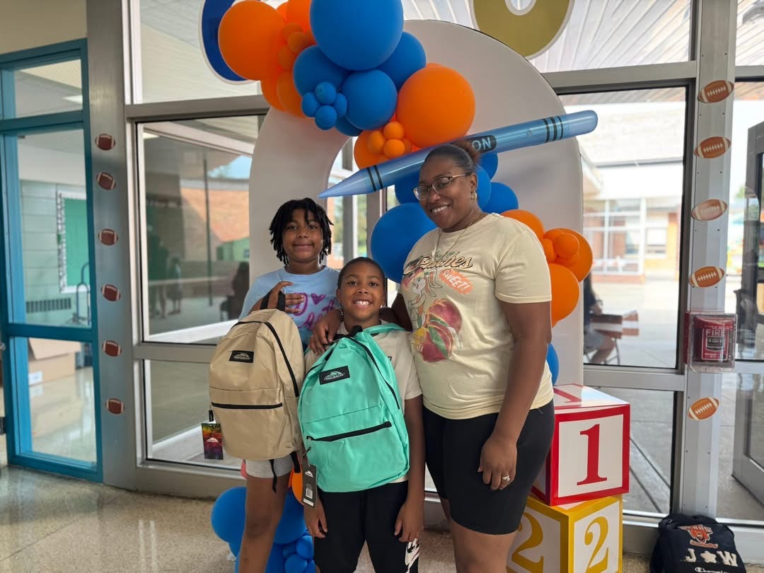 An adult woman and two young children are posing together. The two children are holding up backpacks in front of a colorful back-to-school balloon display.