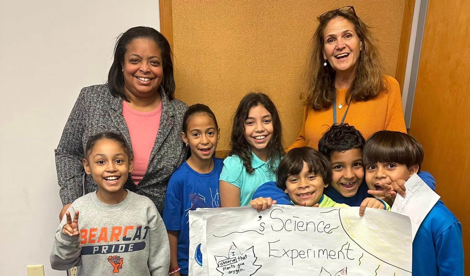 A group of six smiling children hold a large poster labeled “Science Experiment” with colorful drawings and text, standing in front of a brown wall. Two adult women stand behind them, also smiling. The setting appears to be a classroom or school hallway.