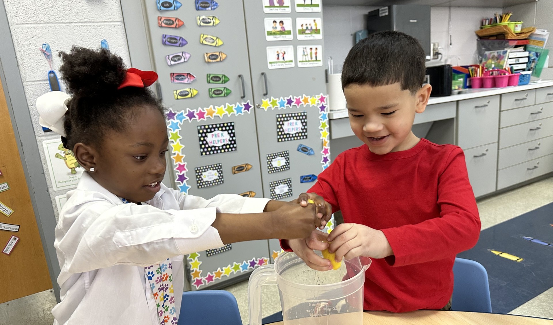Two young students are squeezing lemons into a pitcher in a school classroom setting.