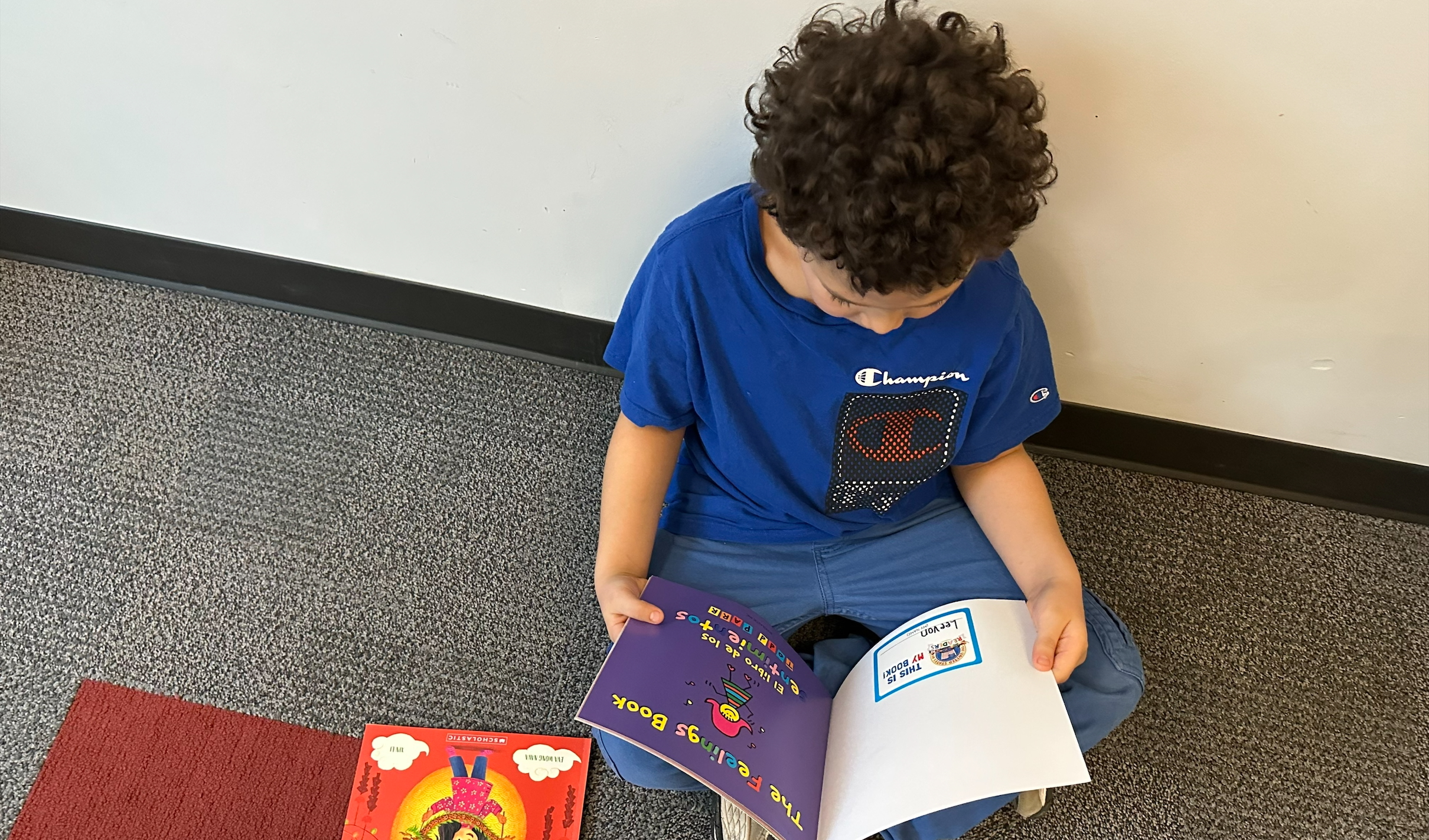 A young child with curly hair, wearing a blue Champion shirt and jeans, sits on gray carpet by a wall, looking at an open book. Two more colorful childrens books, one red and one purple, lie on the floor nearby.