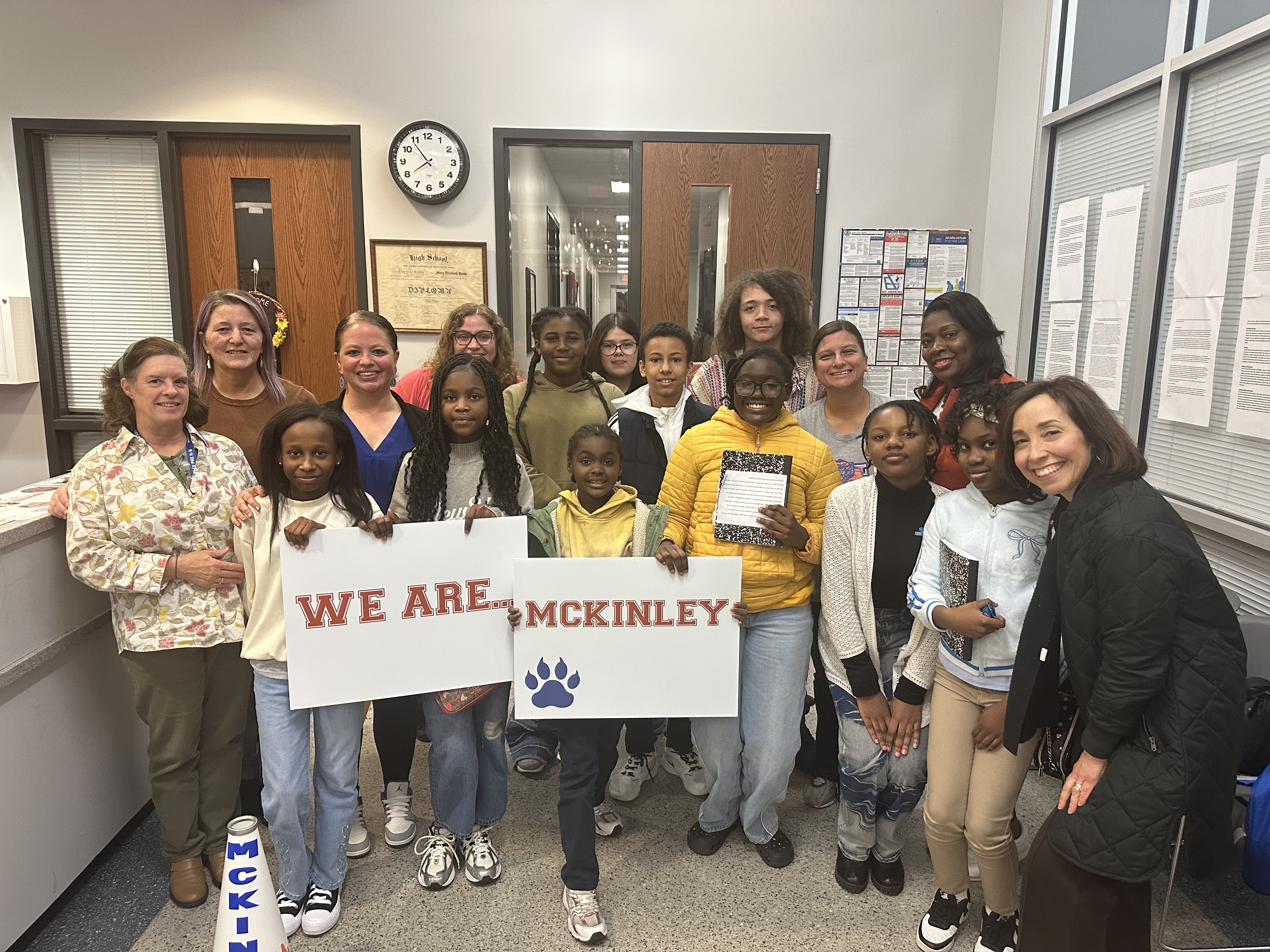 A group of students, educators, and staff members are gathering together holding "We Are... McKinley" signs during a School Board Meeting.
