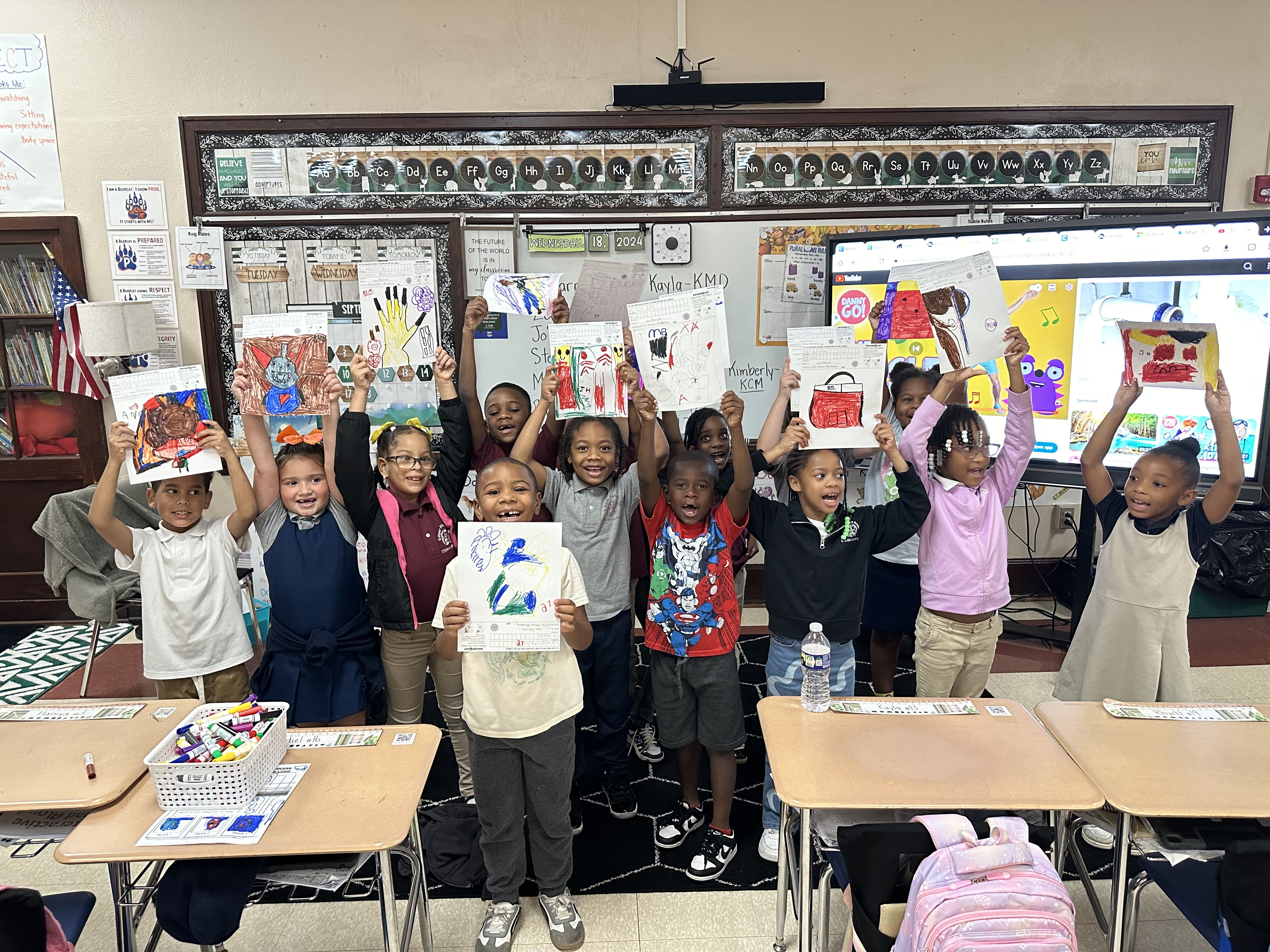 A group of elementary school children smiling and holding up their artwork in a classroom. Desks are arranged around them, with colorful drawings displayed. Educational materials are visible on the wall and a screen in the background.