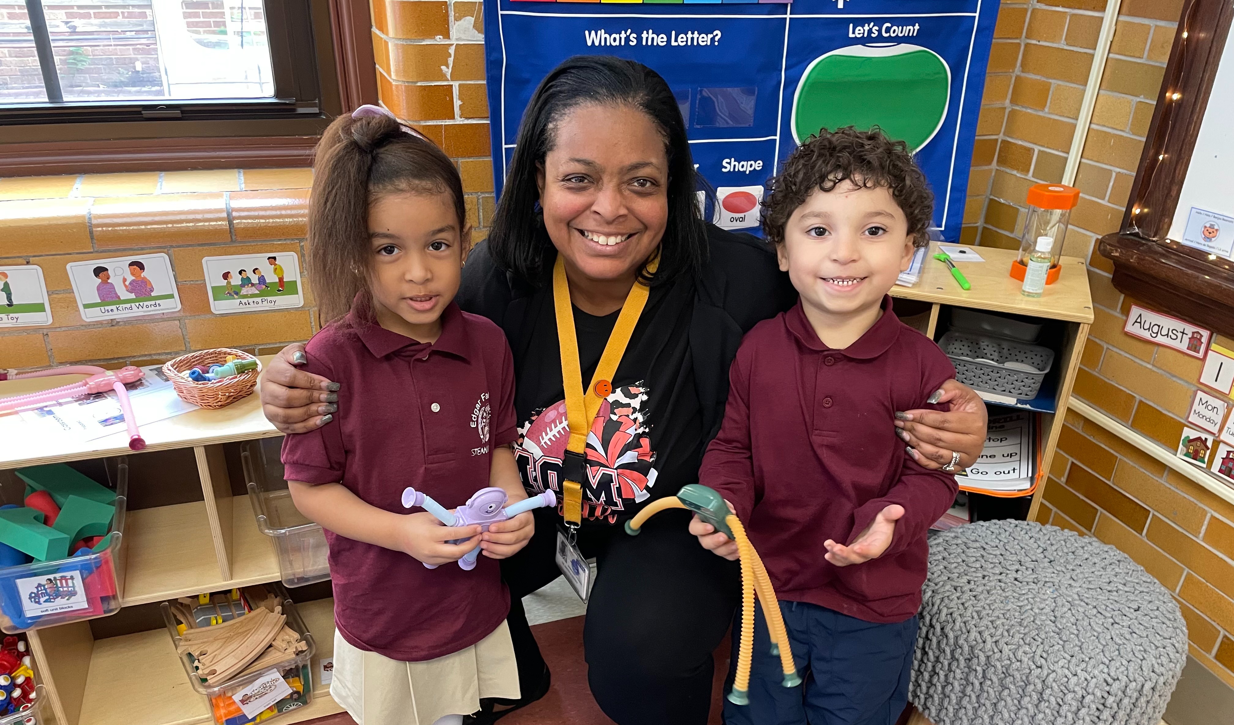 Dr. Andrea Berry-Brown, Superintendent of Schools, is smiling while squatting in the middle of two young students in a school classroom.