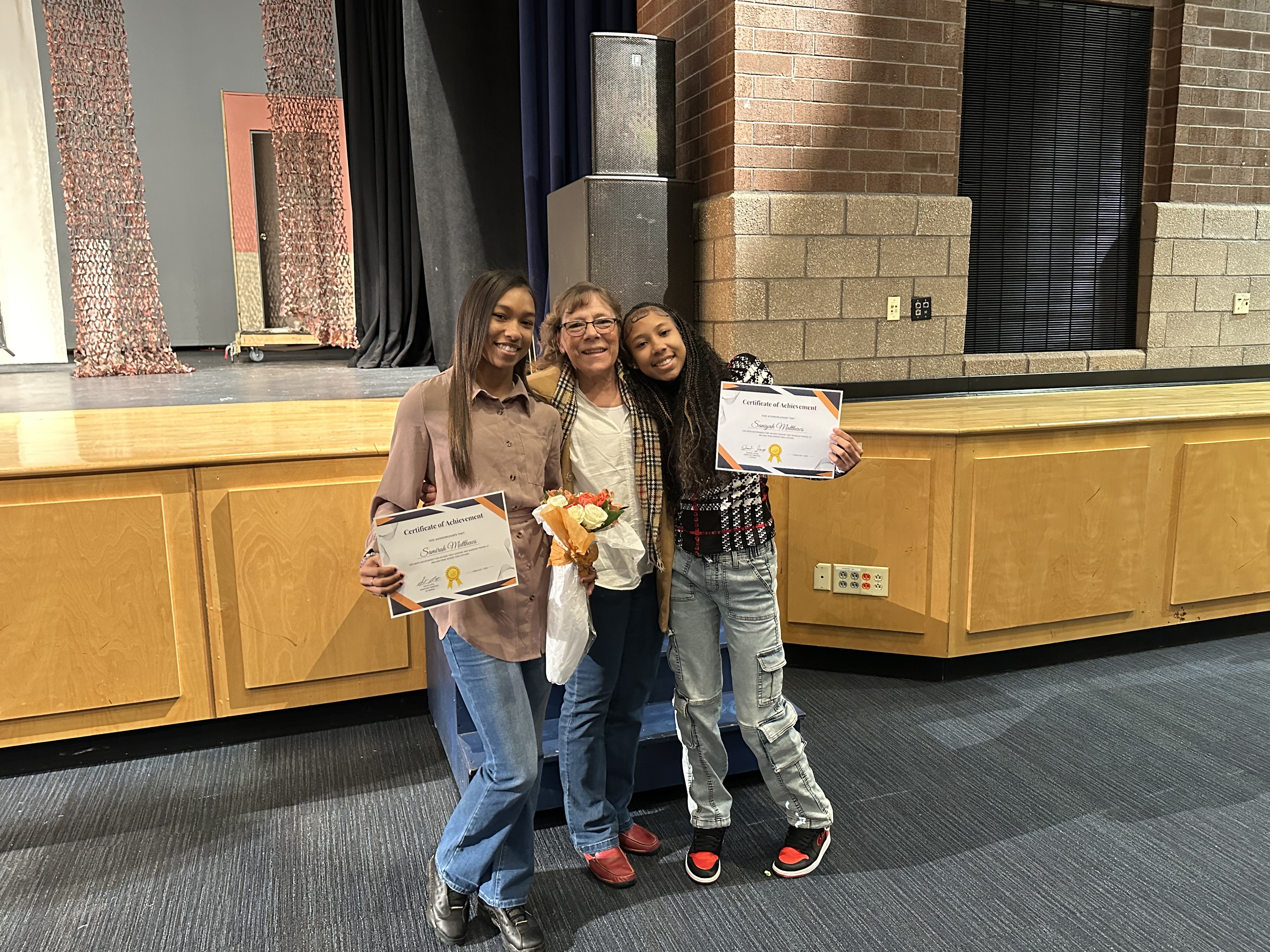 Two young women holding certificates standing beside a woman holding flowers on a stage. The background features a curtain and wooden stage area.