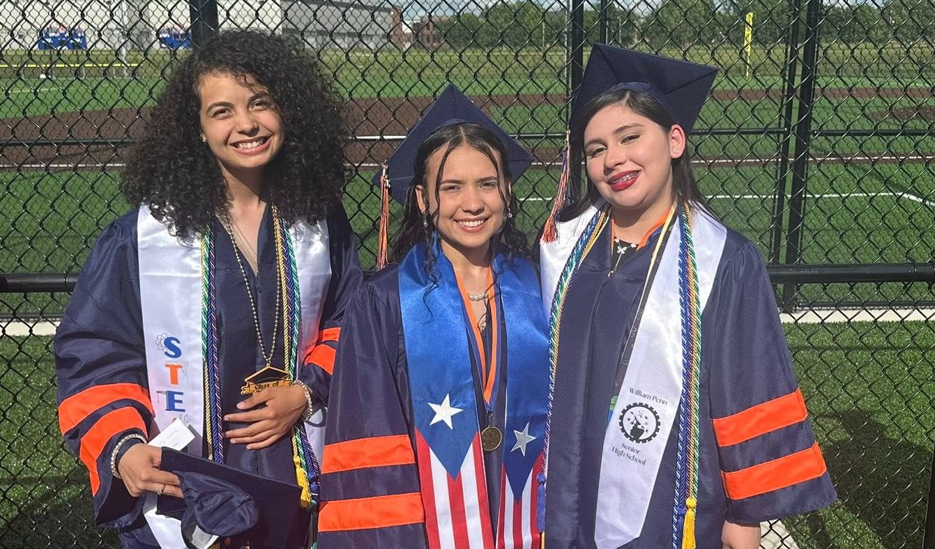 Three students dressed in caps and gowns are posing together outdoors, wearing honor stoles and cords at a graduation ceremony.