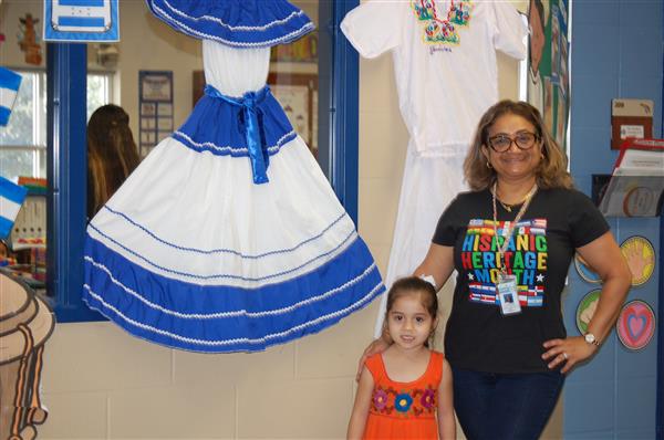 Woman posing with colorful dresses, including a child's blue and white one, in front of a classroom setting.