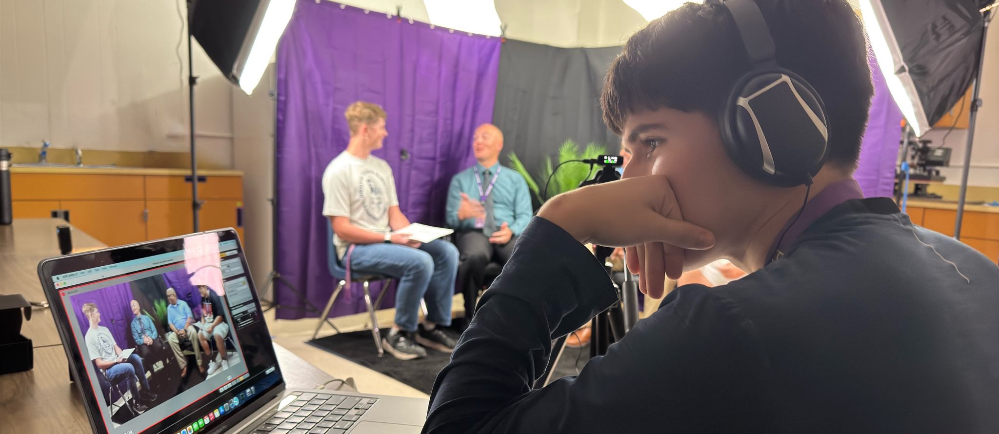 student wearing headphones sitting in front of a podcast session recording monitoring a laptop with video of podcast