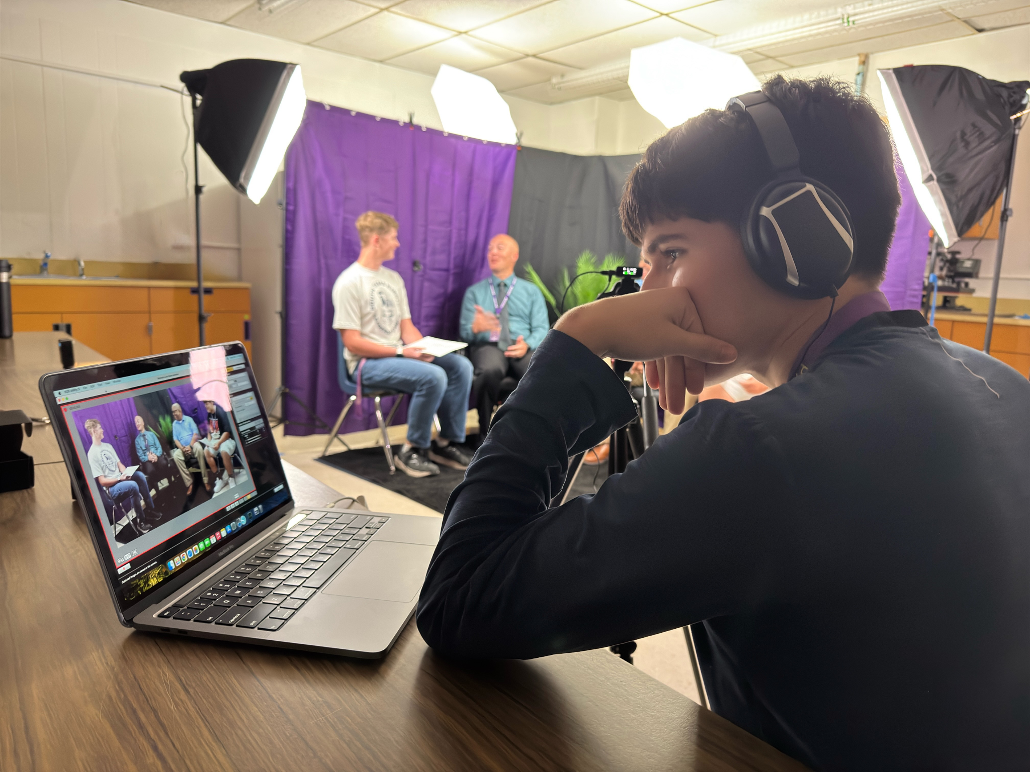 student wearing headphones sitting in front of a podcast session recording monitoring a laptop with video of podcast