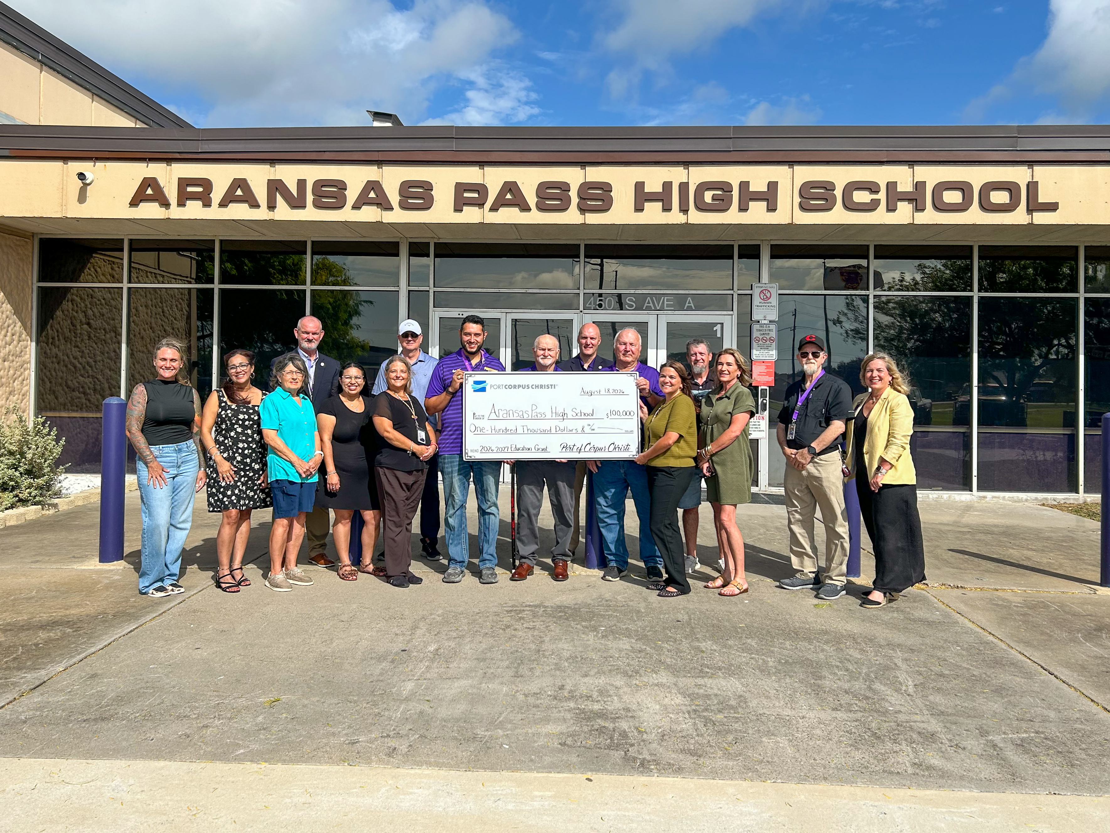 group of people standing in front of a building with the signage "Aransas Pass High School" holding a ceremonial check.