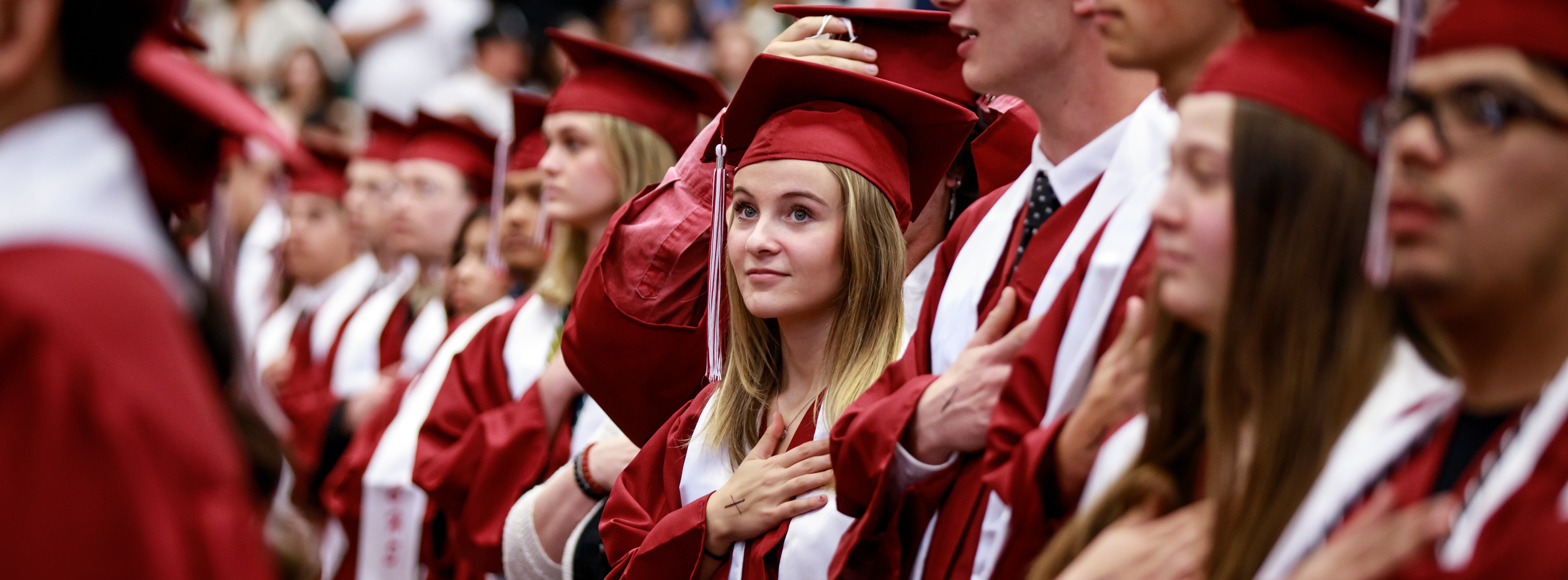 A Columbus High School senior holds her hand over her heart during the playing of the National Anthem during the 2024 CHS Commencement ceremony. She is centered in crowd of other graduates all wearing the same maroon cap and gown.