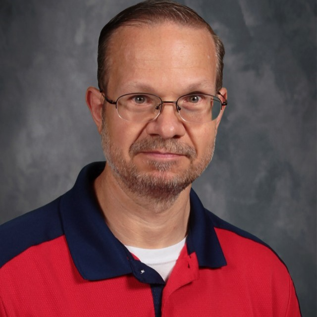 =Brian Townsend, poses for a photo in a red shirt with blue collar. He has short brown hair, mixed gray beard  and is wearing thin-rimmed glasses.