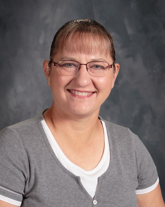 A women wearing a gray sweater and blacked rimmed glasses poses in front of a black and gray background.