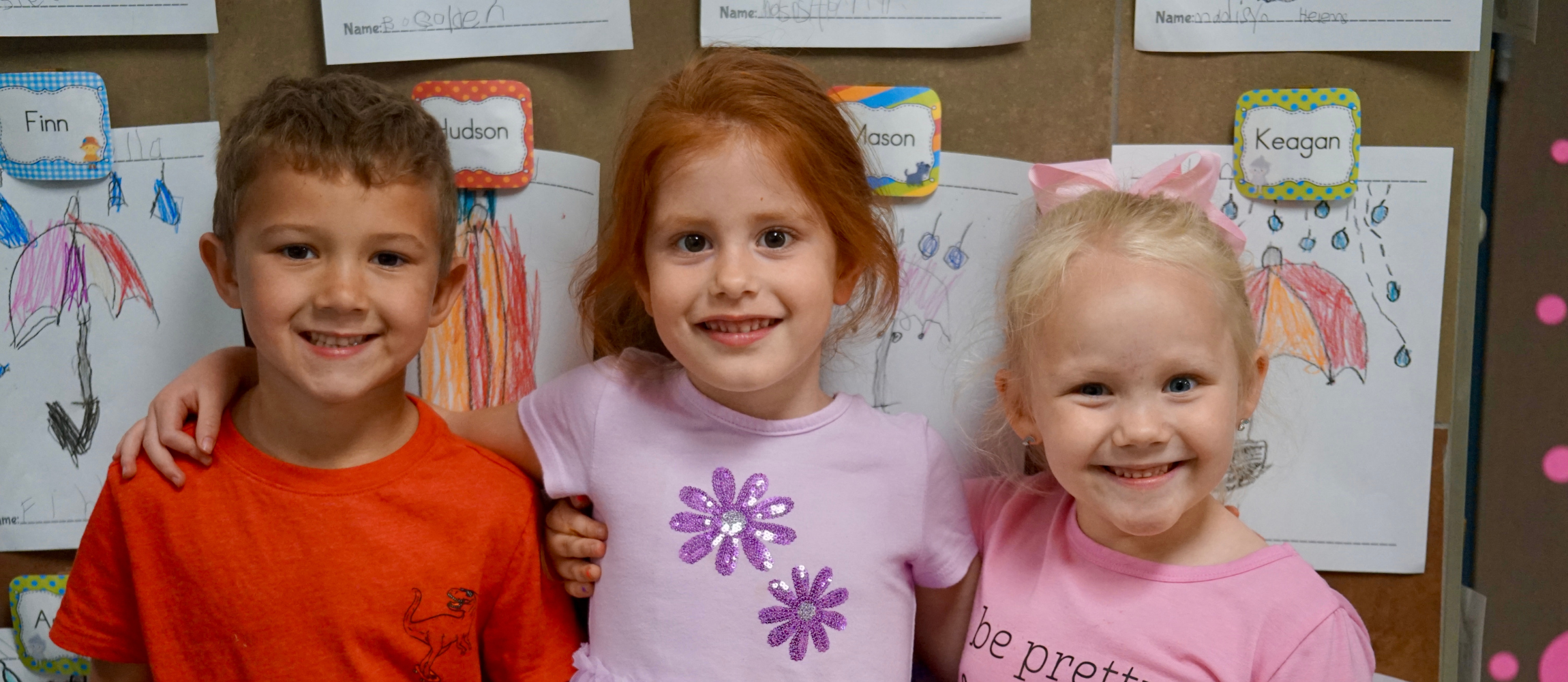 three students standing in classroom smile with each other