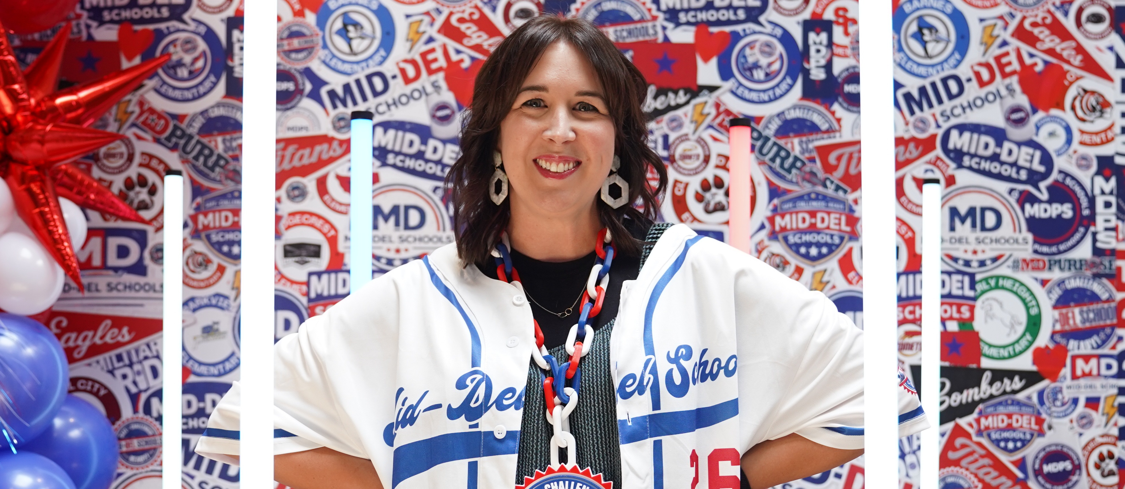 A smiling Mid-Del Schools team member stands with hands on her hips in front of a colorful Mid-Del Schools backdrop covered with school logos and district branding. She is wearing a white and blue Mid-Del baseball-style jersey, a red, white, and blue chain necklace with a "Game Changer Ready" medallion, and white geometric earrings. Red, white, and blue balloons and illuminated light pillars frame the photo, creating a fun, energetic atmosphere that celebrates school pride and the Mid-Del Experience.