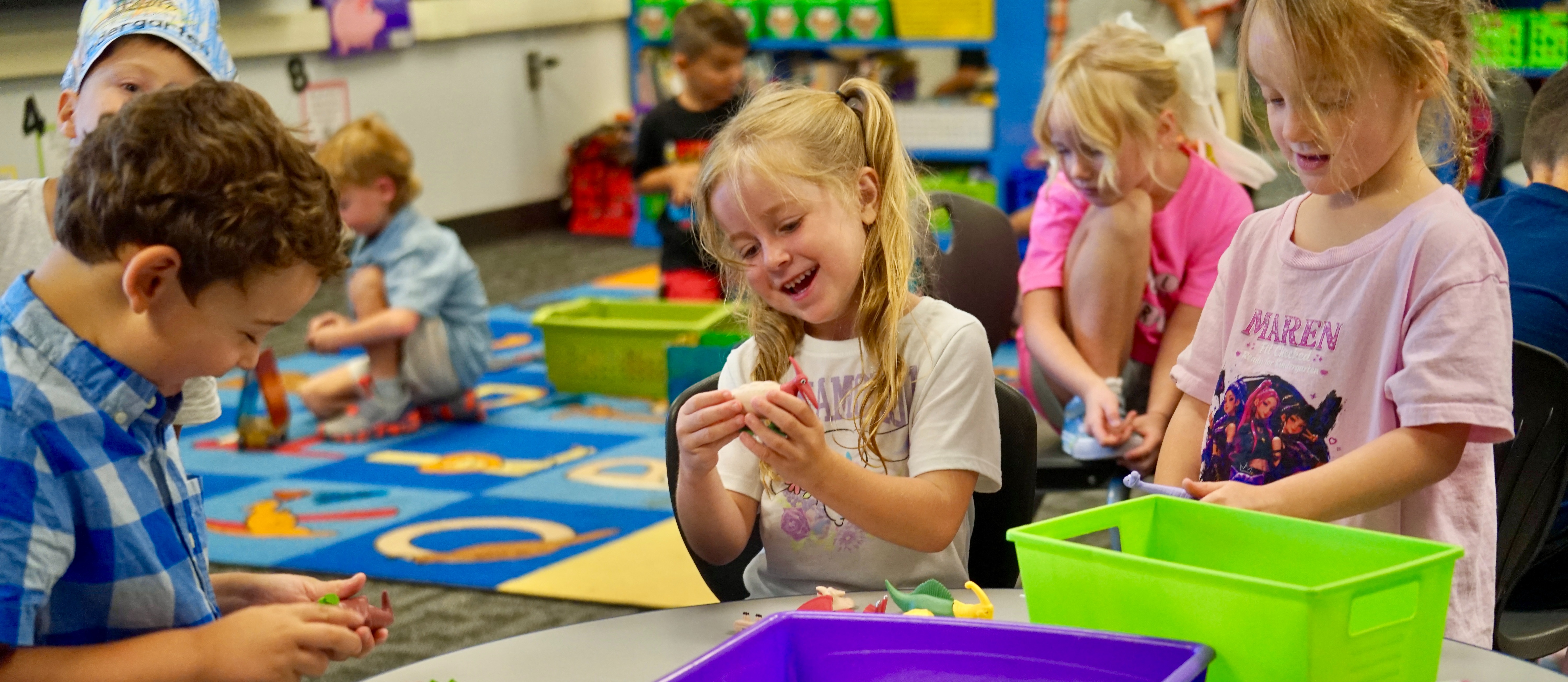 Several young children sit around a table in a classroom, engaging in activities with colorful materials.