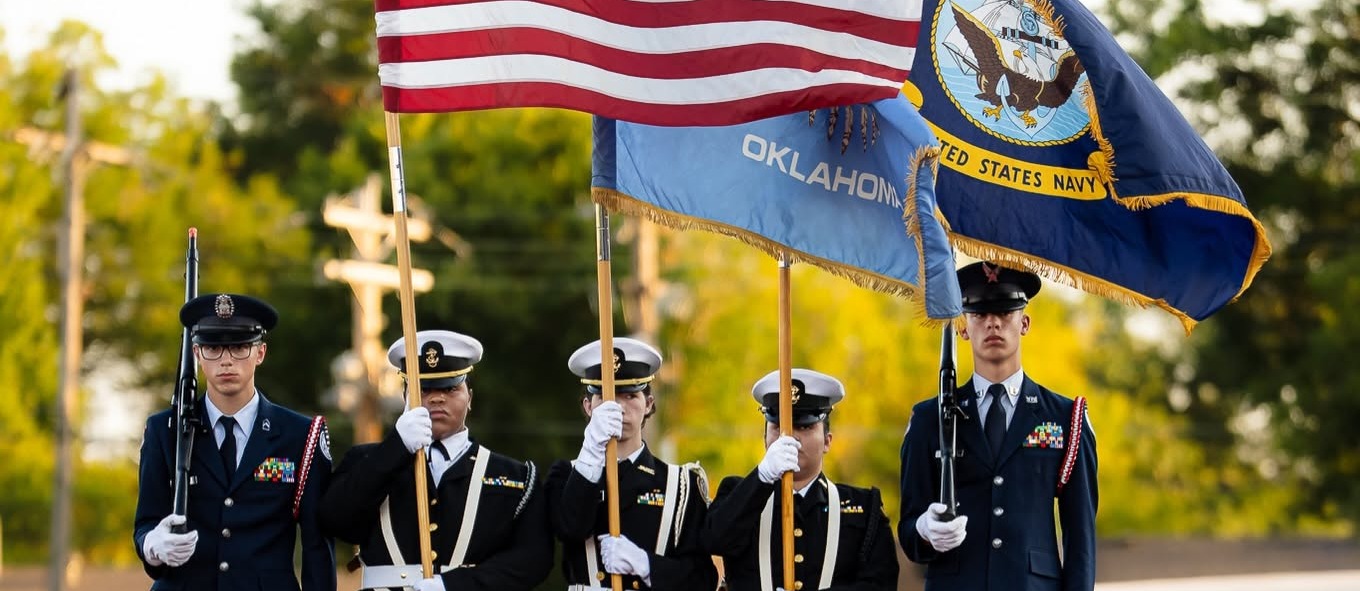 Five uniformed military personnel holding flags on a red carpet. Background shows trees and a building.