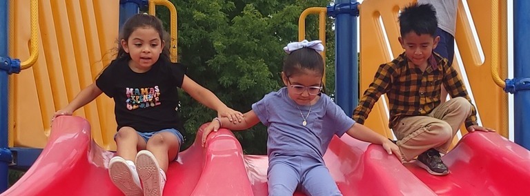 Three young North Park students hold hands while they go down a big pink slide together.