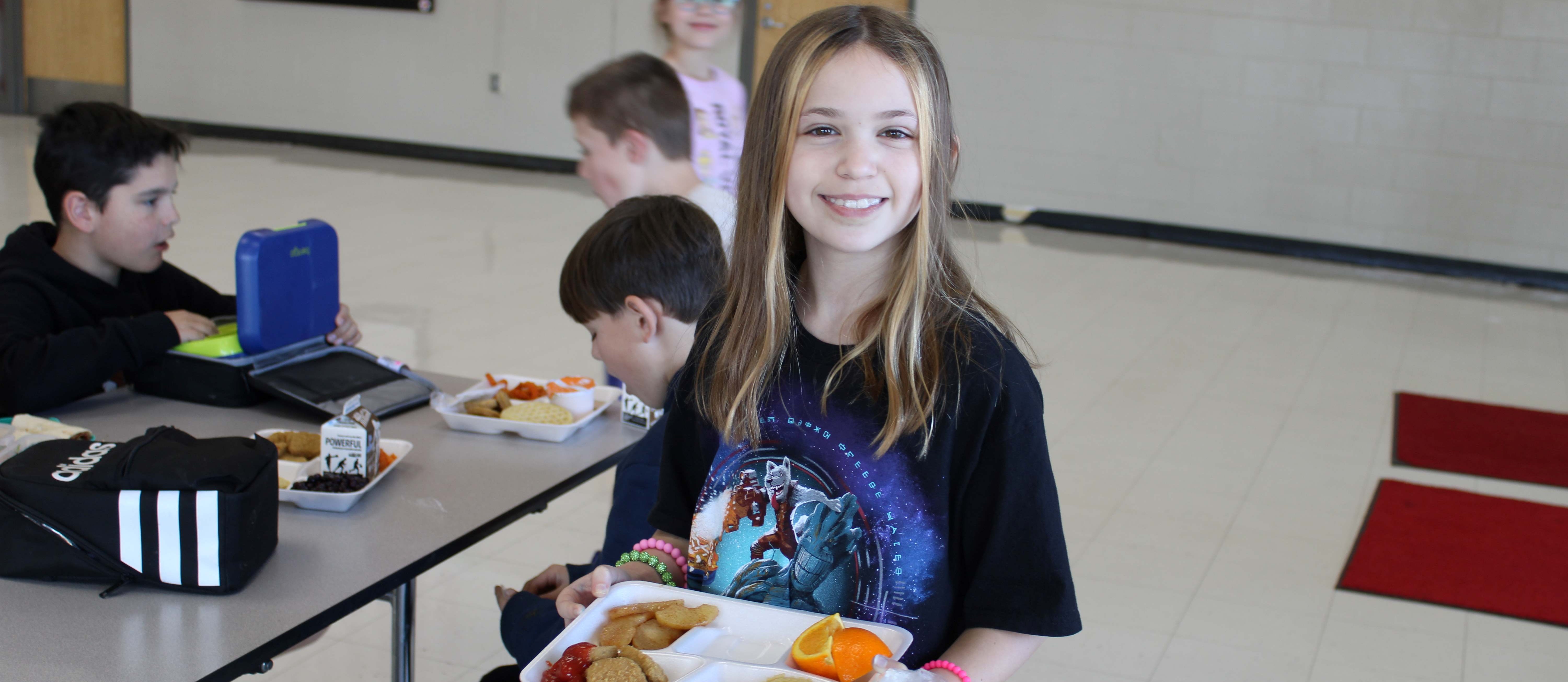 Girl carrying lunch tray in cafeteria. Three students sitting at table eating lunch.