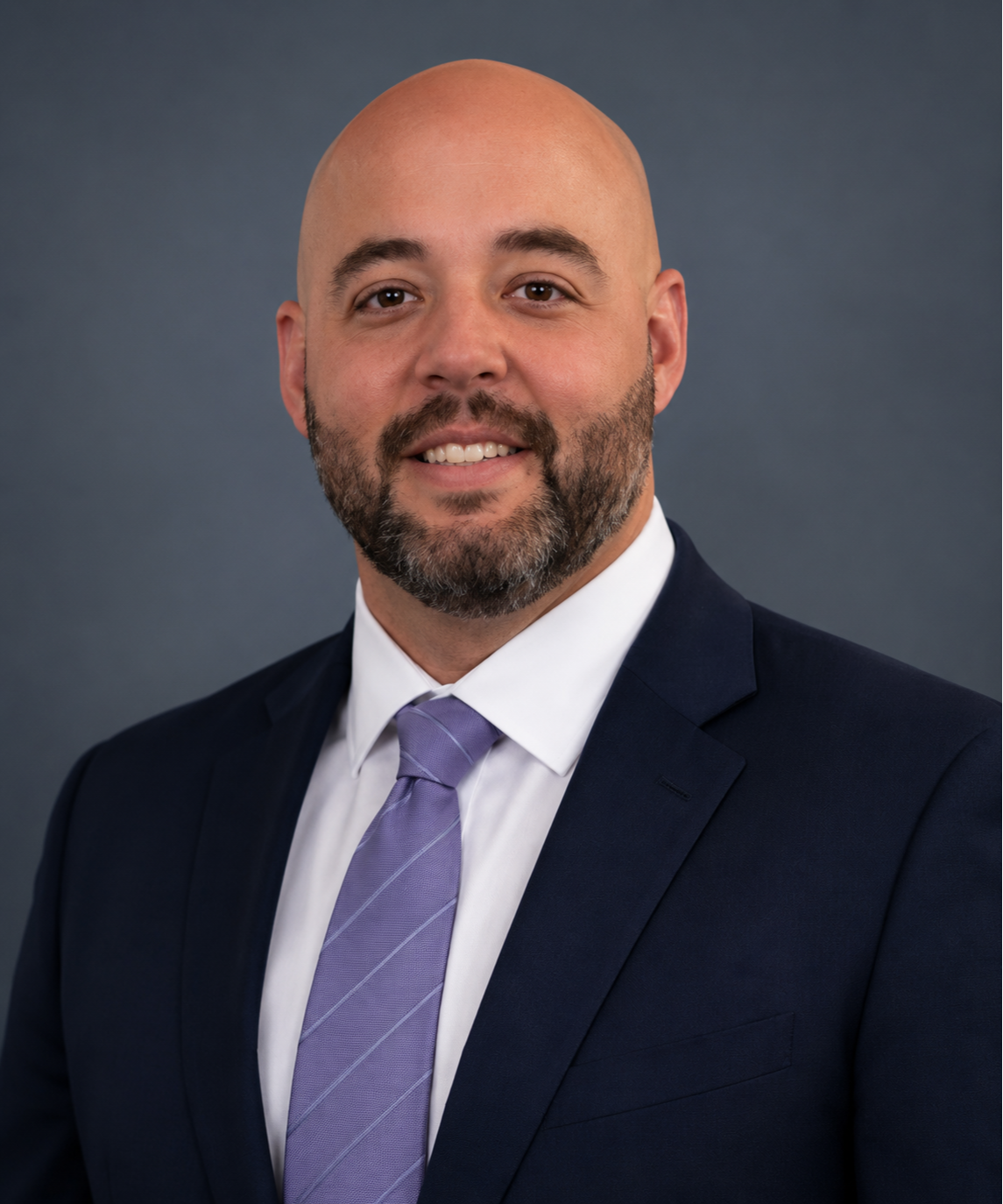 Person in navy suit with purple tie, white collared shirt, and smiling face, with light grey background.
