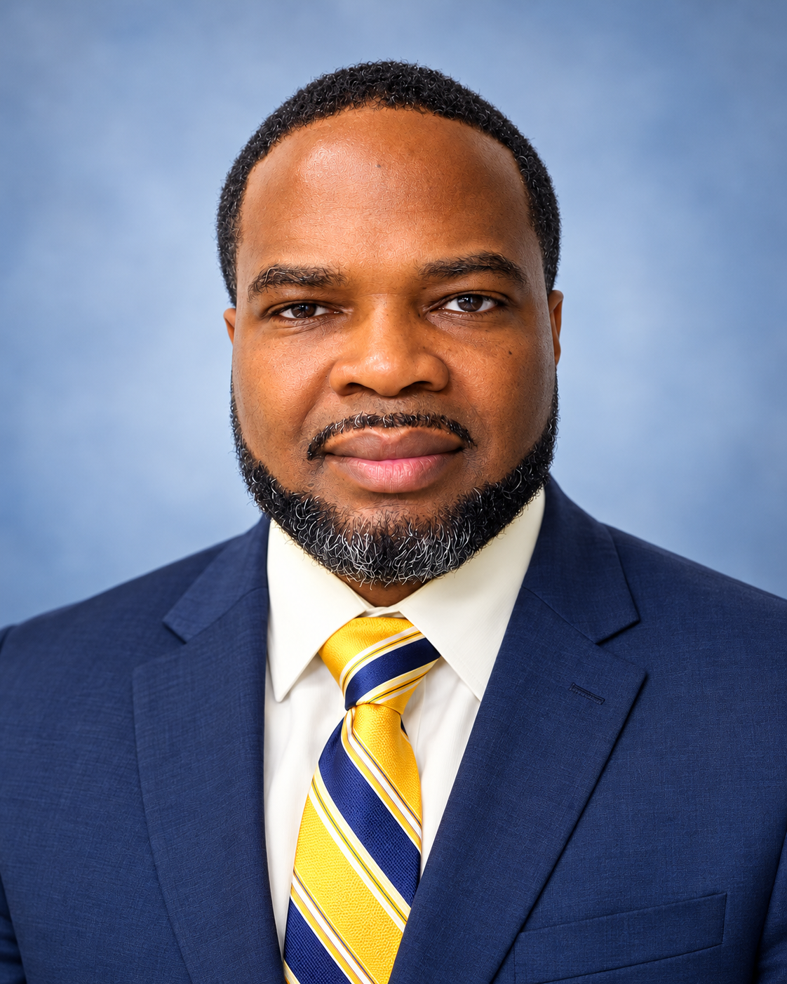 Man in a navy blue suit, white shirt, and yellow tie with stripes. He has a beard and is smiling.