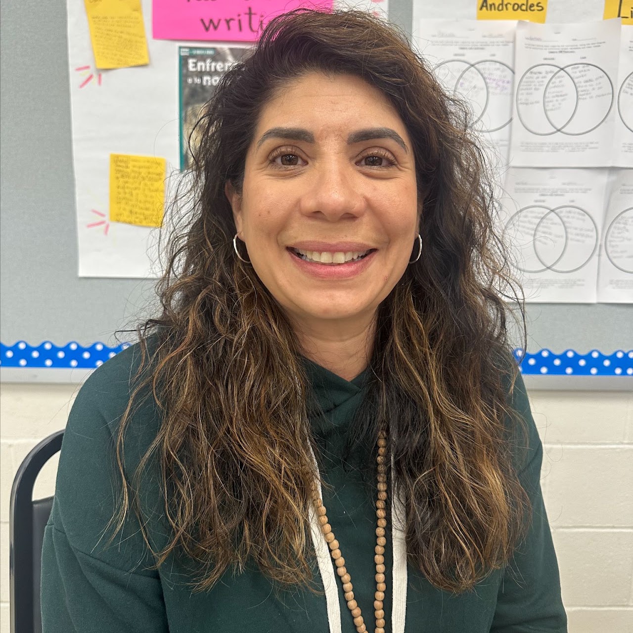 Woman with curly hair, wearing a green shirt, smiling at the camera. She is in a classroom with a bulletin board in the background.