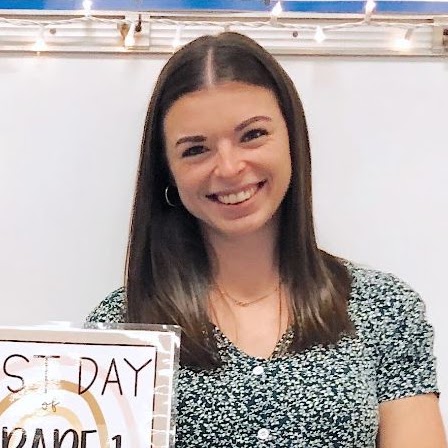 Woman in floral top smiling at camera with sign reading "First Day Grade 1".