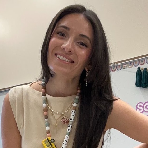 A woman wearing a light-colored blouse and necklace stands in front of a white board with a tasseled banner.
