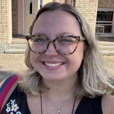Woman with blonde hair, wearing glasses, smiling, standing in front of a building.