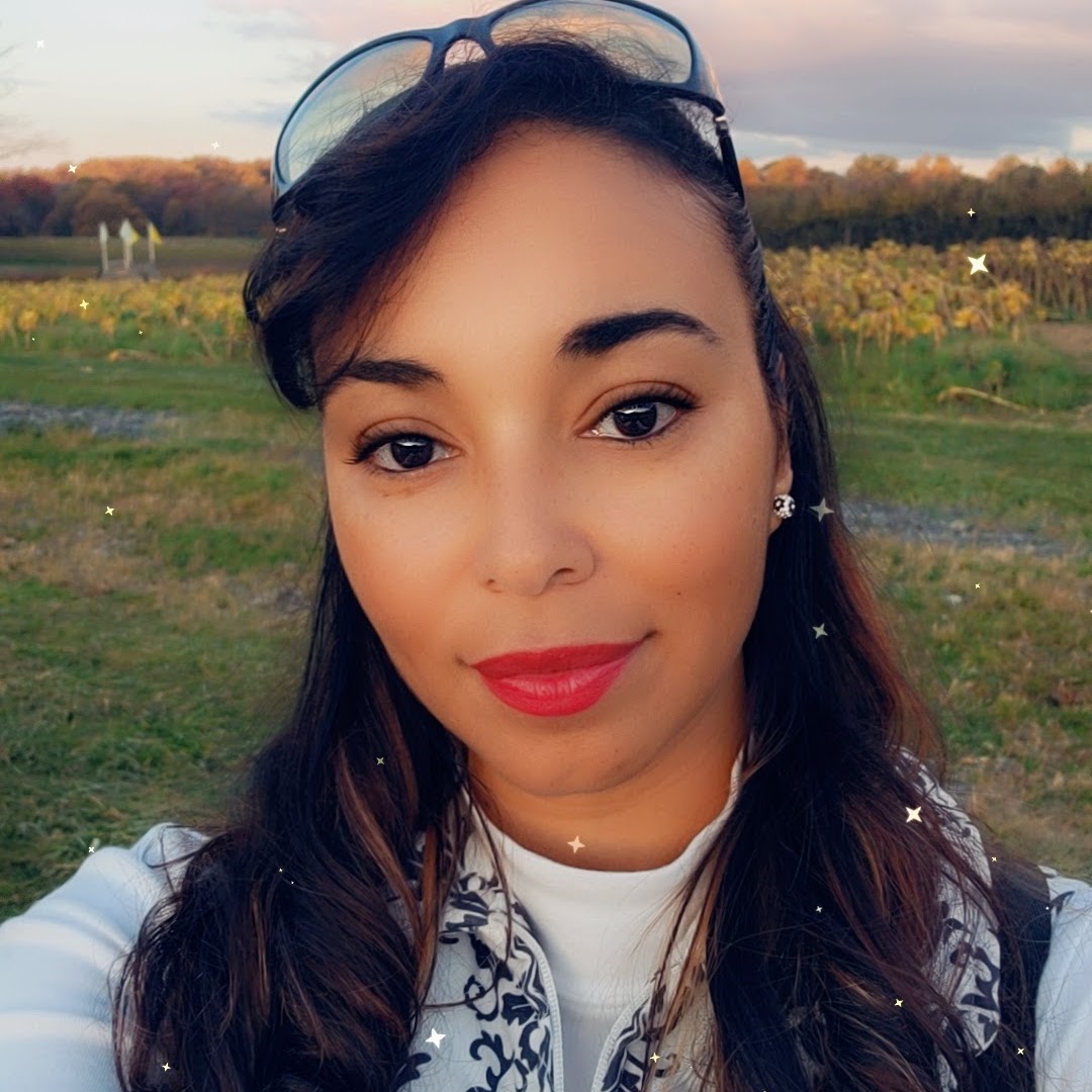 A smiling woman with long, wavy hair and red lipstick takes a selfie outdoors. She wears sunglasses and a white jacket. Behind her is a grassy field with trees.