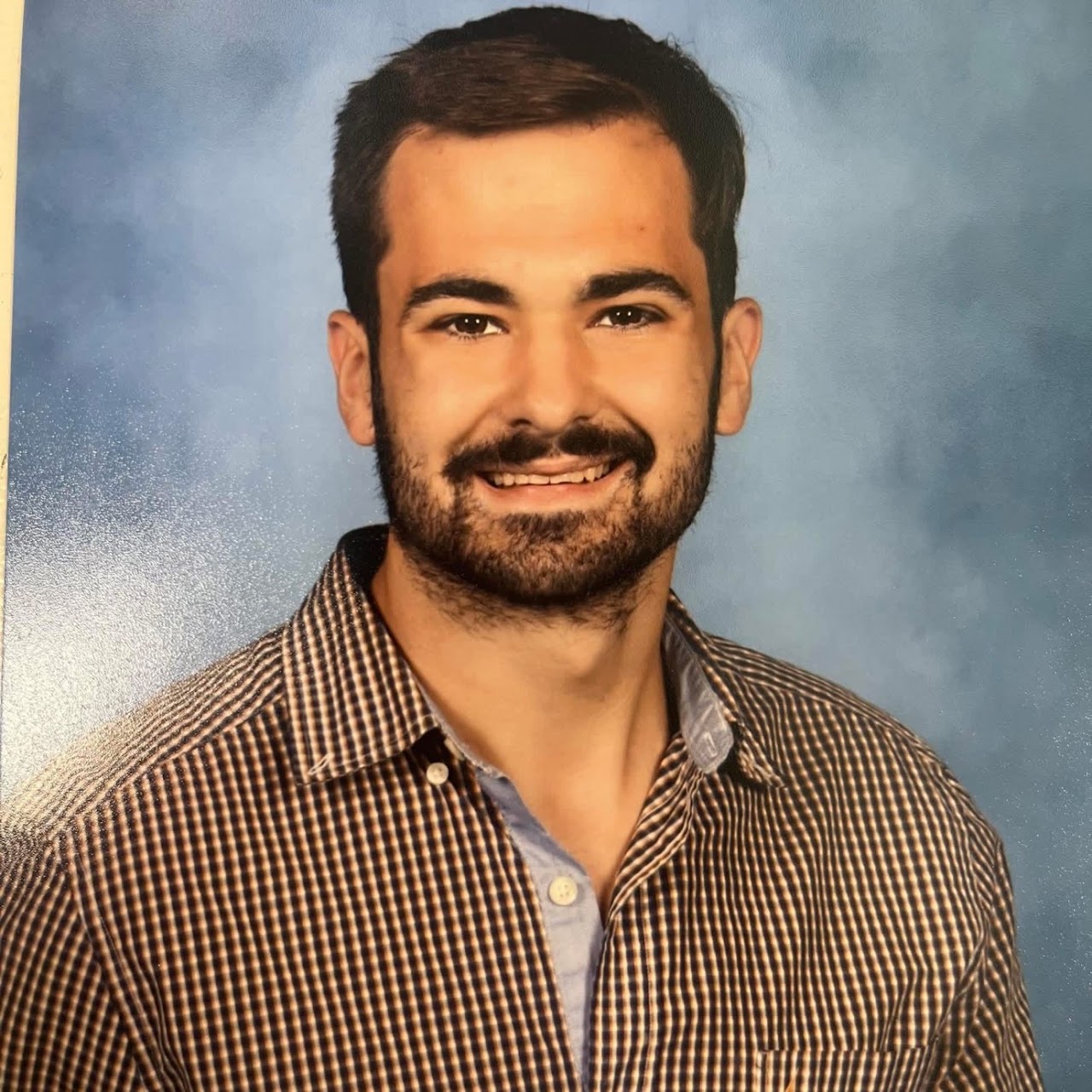 A man with a beard, wearing a brown and white checkered shirt, smiling for a photo.